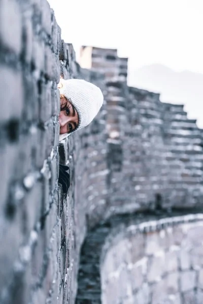 Woman in white winter hat peeking around a large stone wall