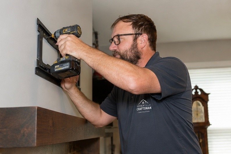 A man installing a wall-mounted TV mount using a cordless drill in a living room.