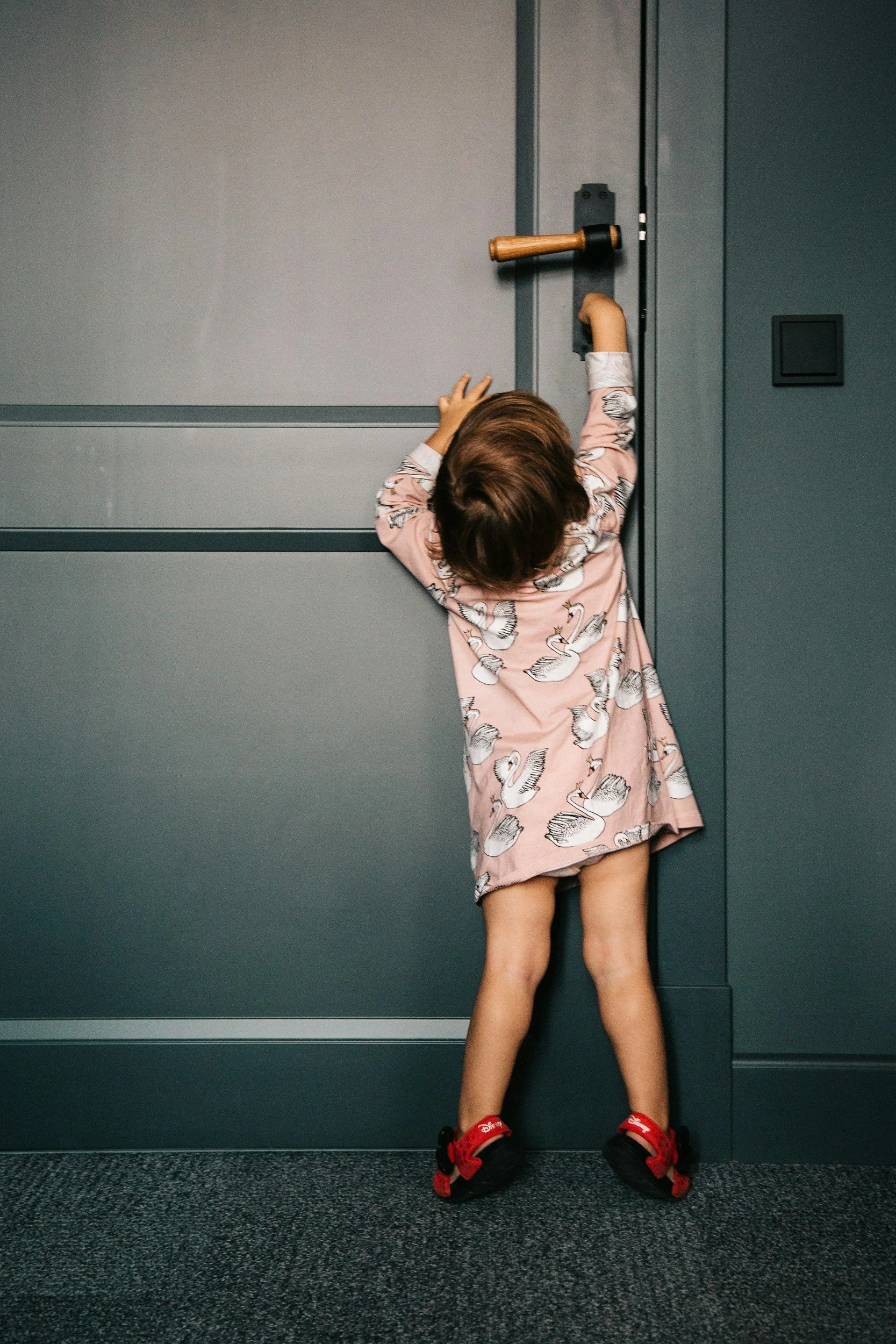 A young child in a pink dress with swan patterns, standing in front of a green door, reaching up to try to open the door latch. Symbolizing on this site that we provide baby proofing for door handles.
