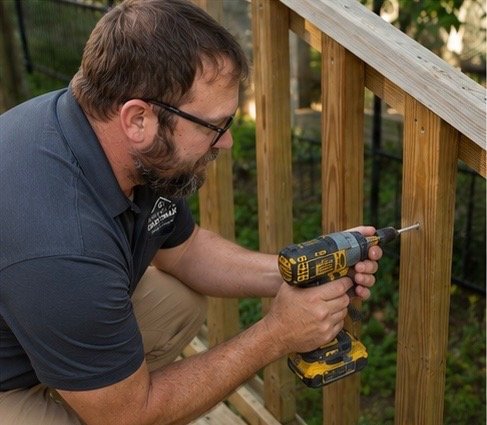 A man using a cordless drill to screw into a wood railing on a deck in a backyard. Symbolizing deck repairs.