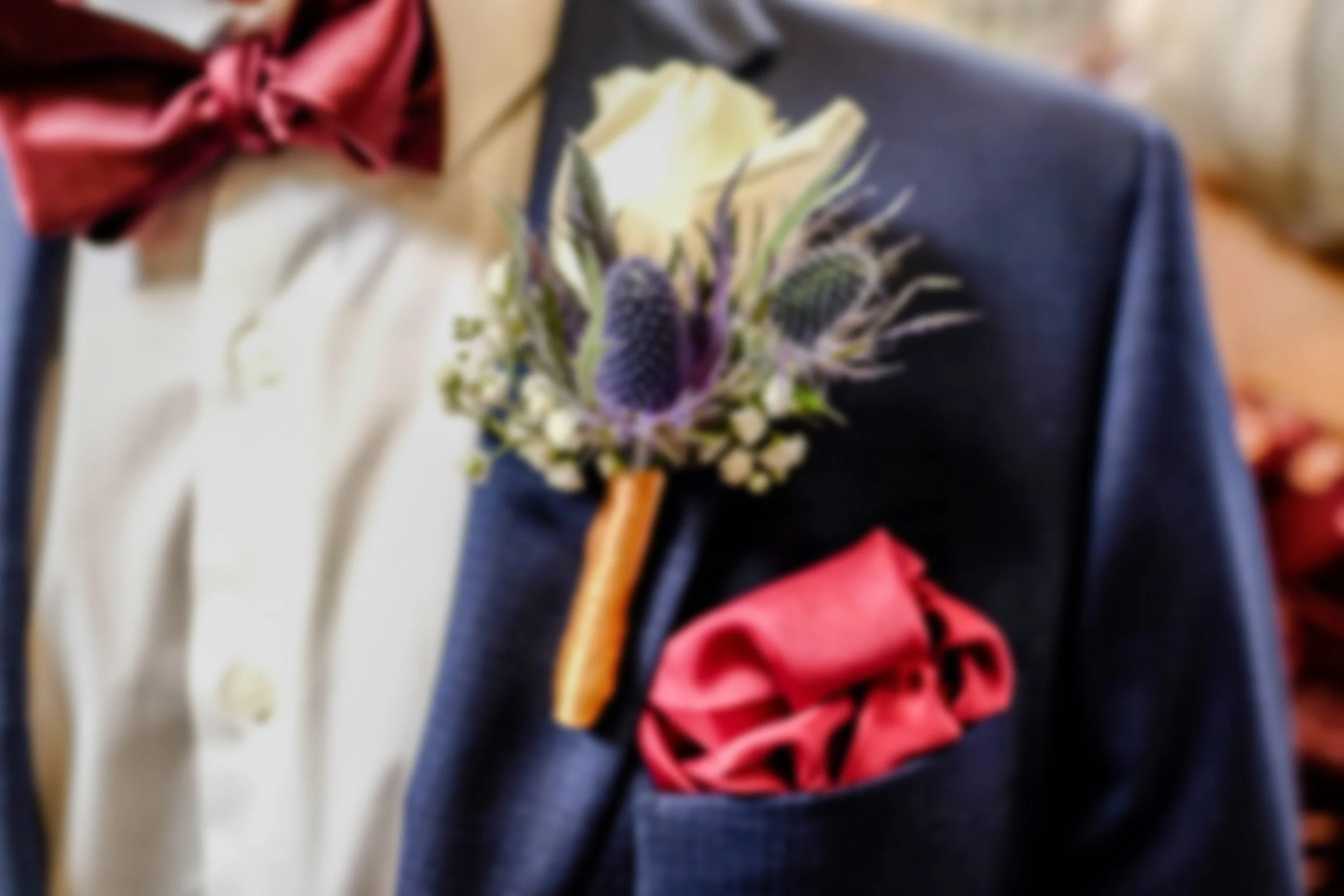 Close-up of a person wearing a tuxedo with a white shirt, navy jacket, maroon bow tie, and a boutonniere with a white rose and blue accents.