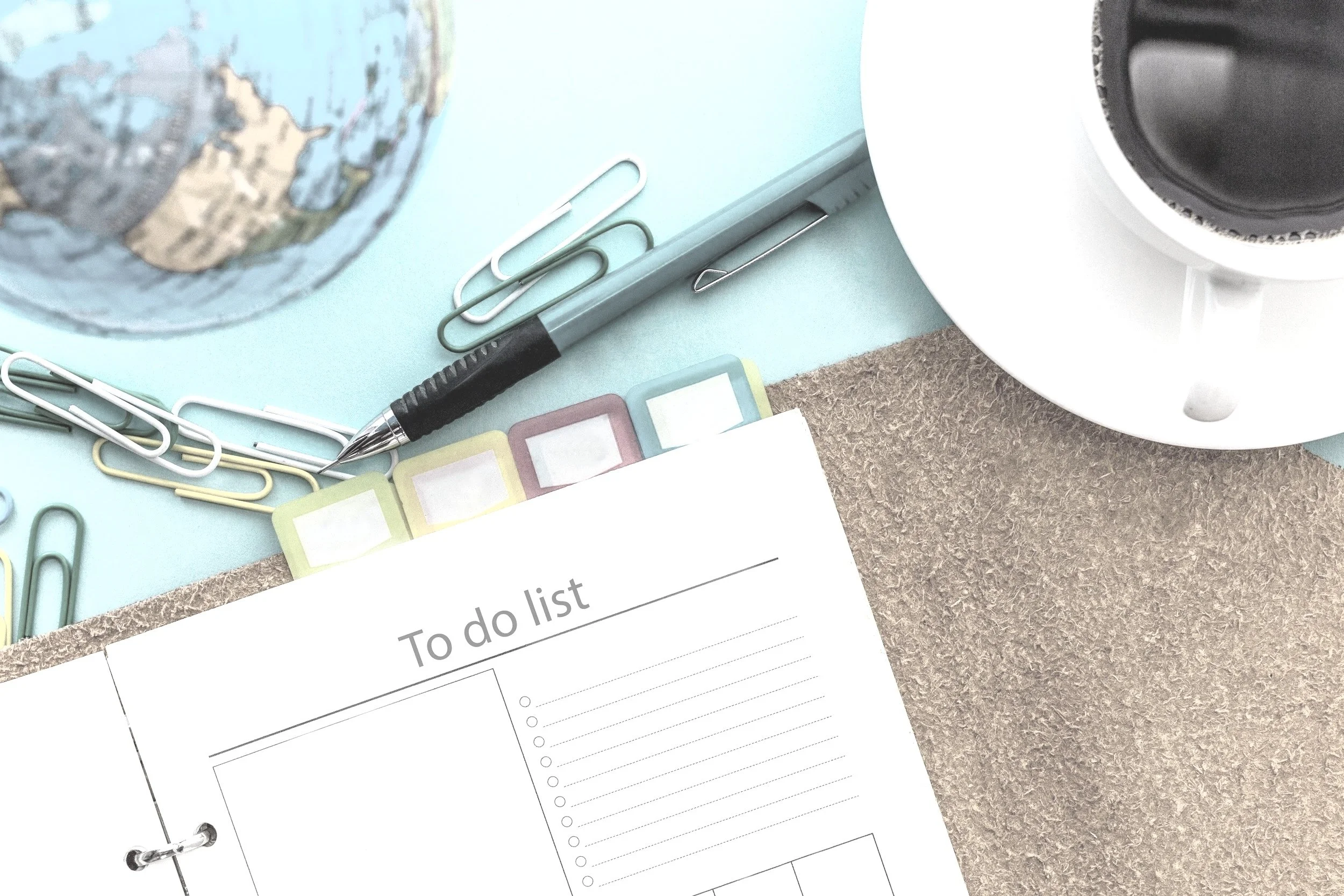 A flat lay of a desk with a world globe, paper clips, a black pen, sticky notes, a to-do list planner, and a partial view of a white electronic device, possibly a speaker or humidifier.