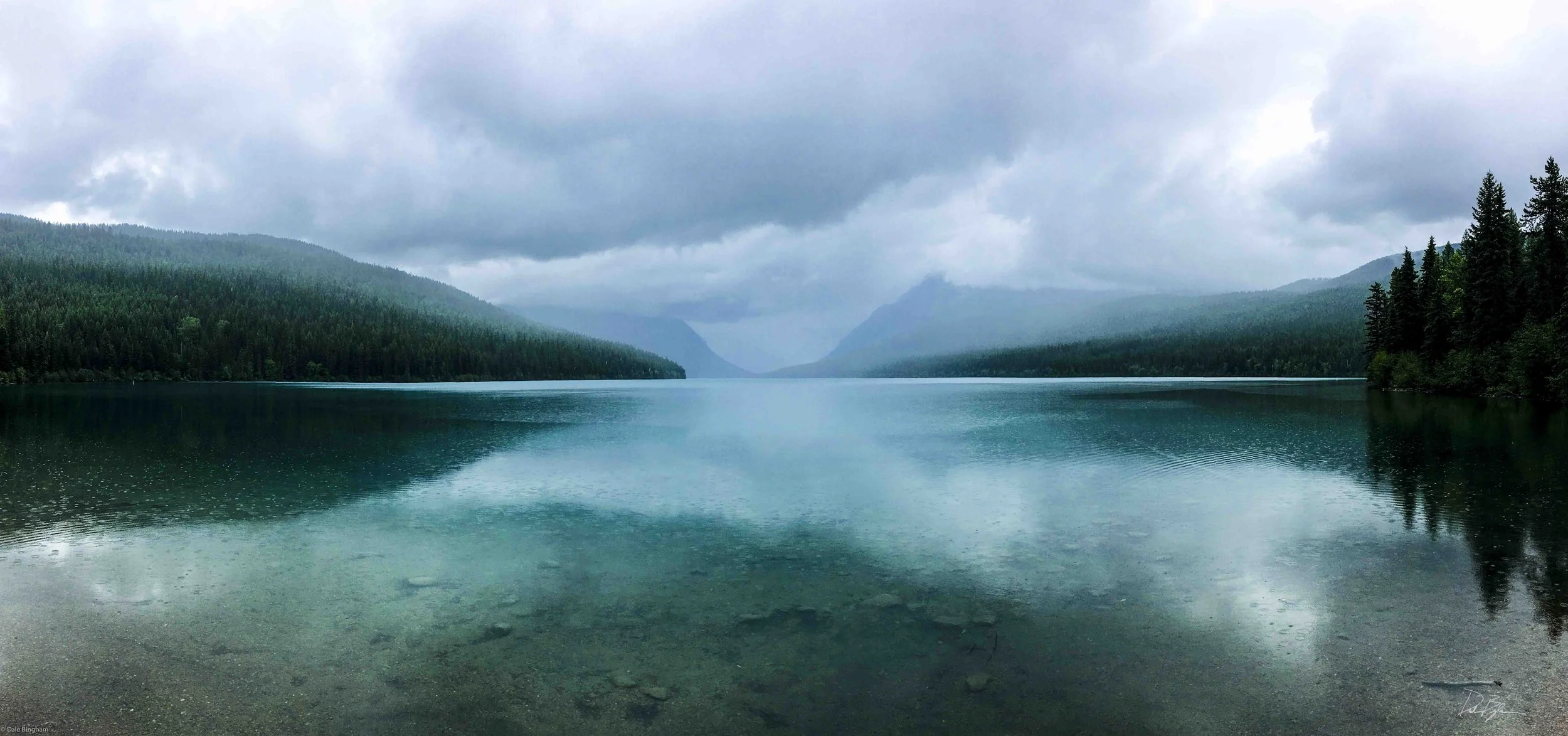 Calm lake surrounded by forested mountains under cloudy sky.