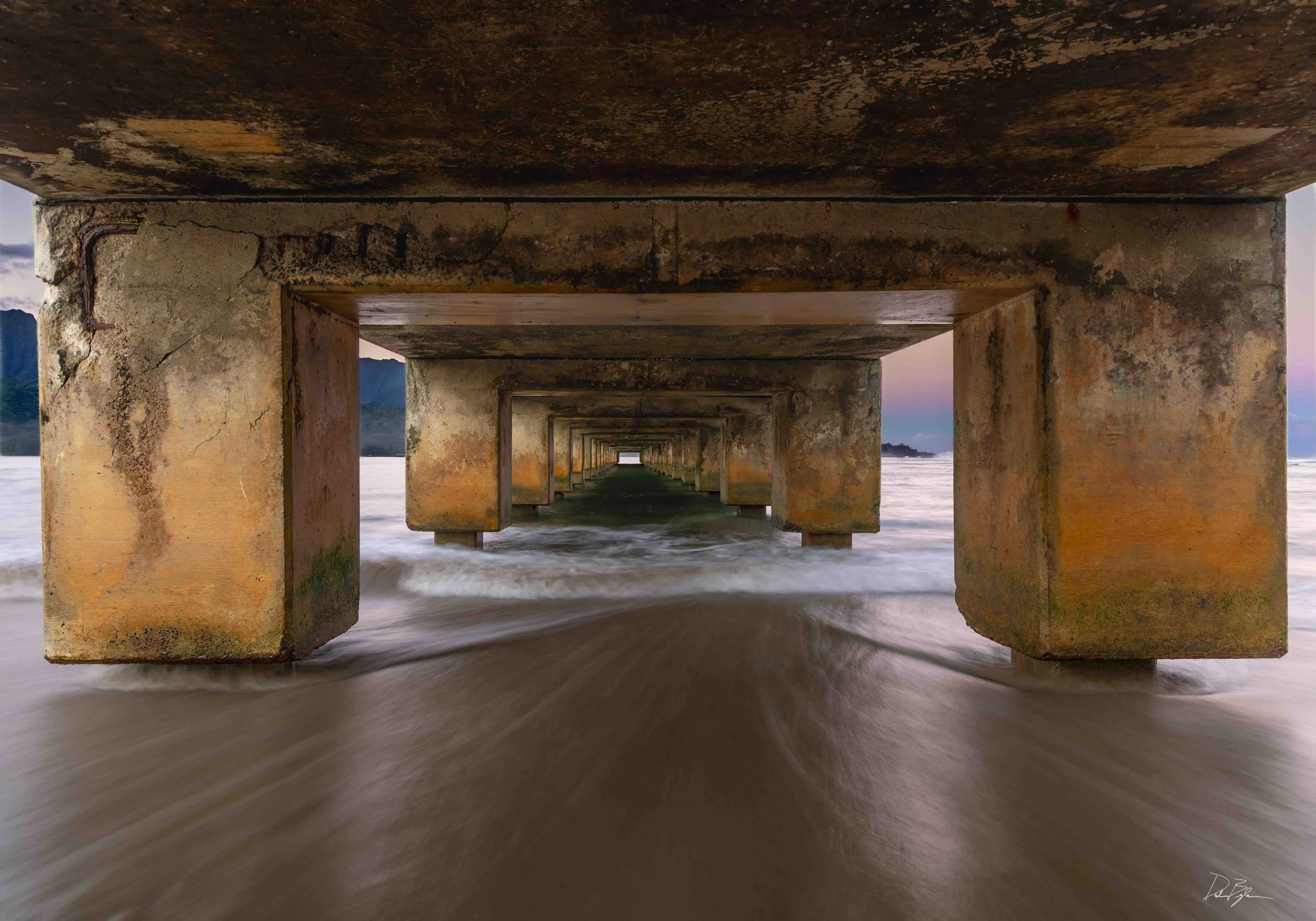 View underneath a weathered pier extending into the water during sunset with mountain background. Kauai photography.  Hanalei Bay photography.