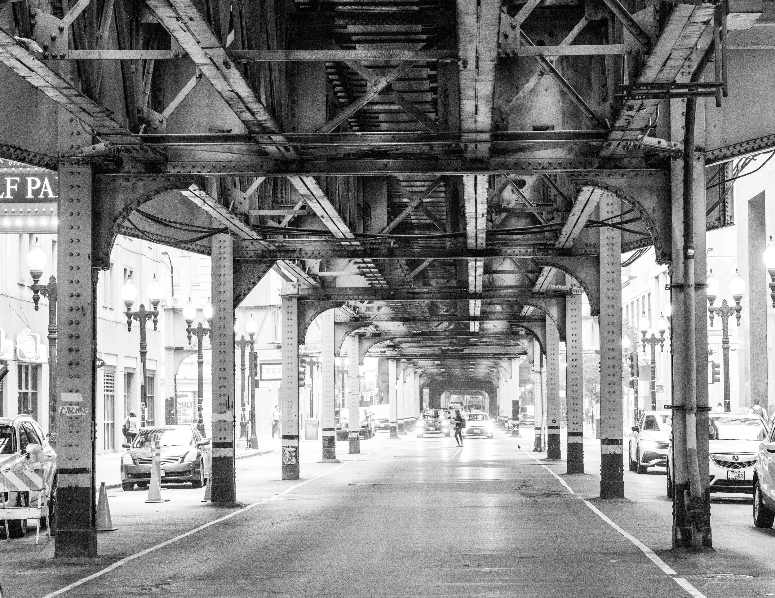 Black and white photograph of an elevated train track structure in an urban area, with cars on the street below and city buildings on either side.