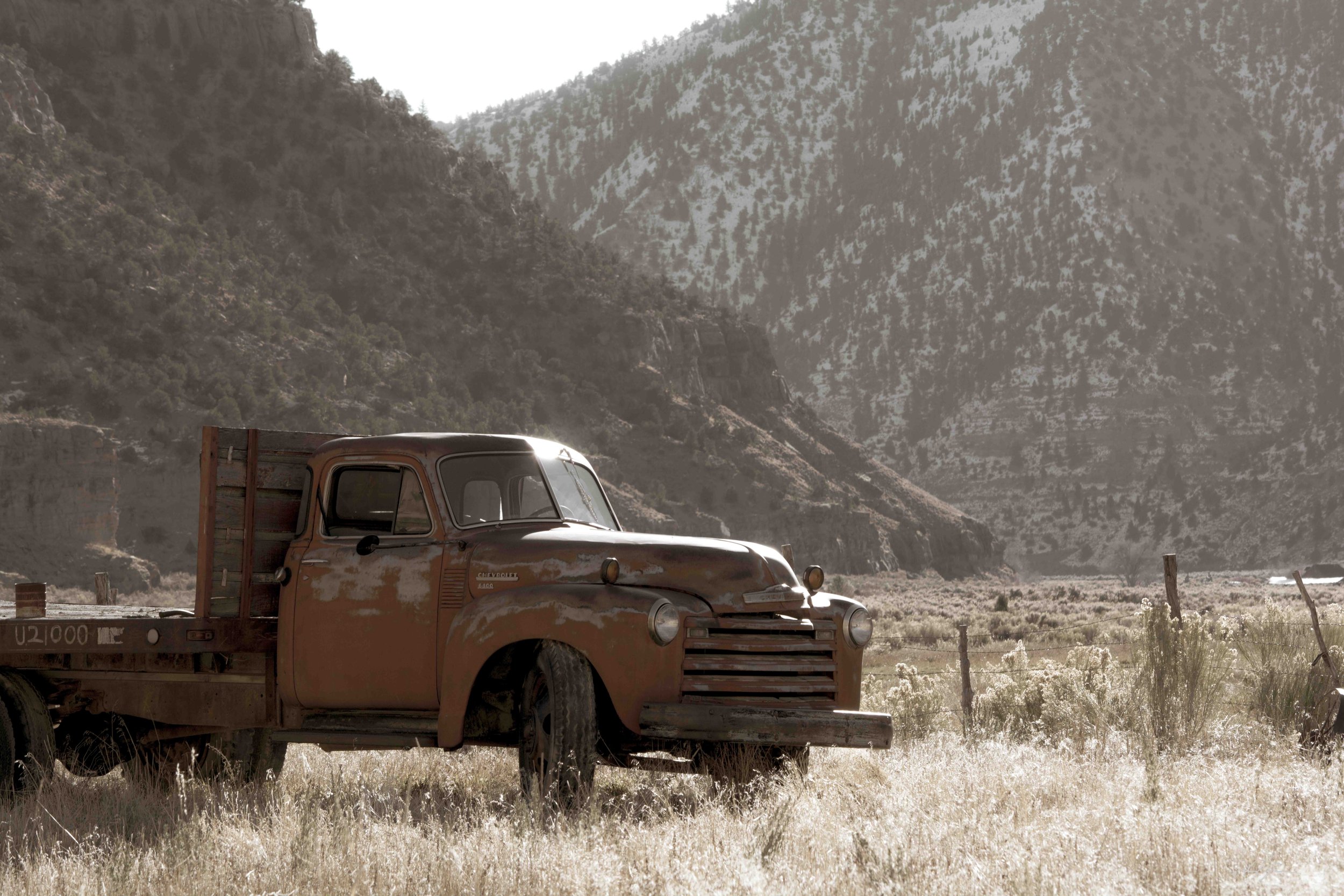An old, rusty orange Chevrolet truck parked in a grassy field near a wooden fence, with snow-covered mountains in the background under a cloudy sky.