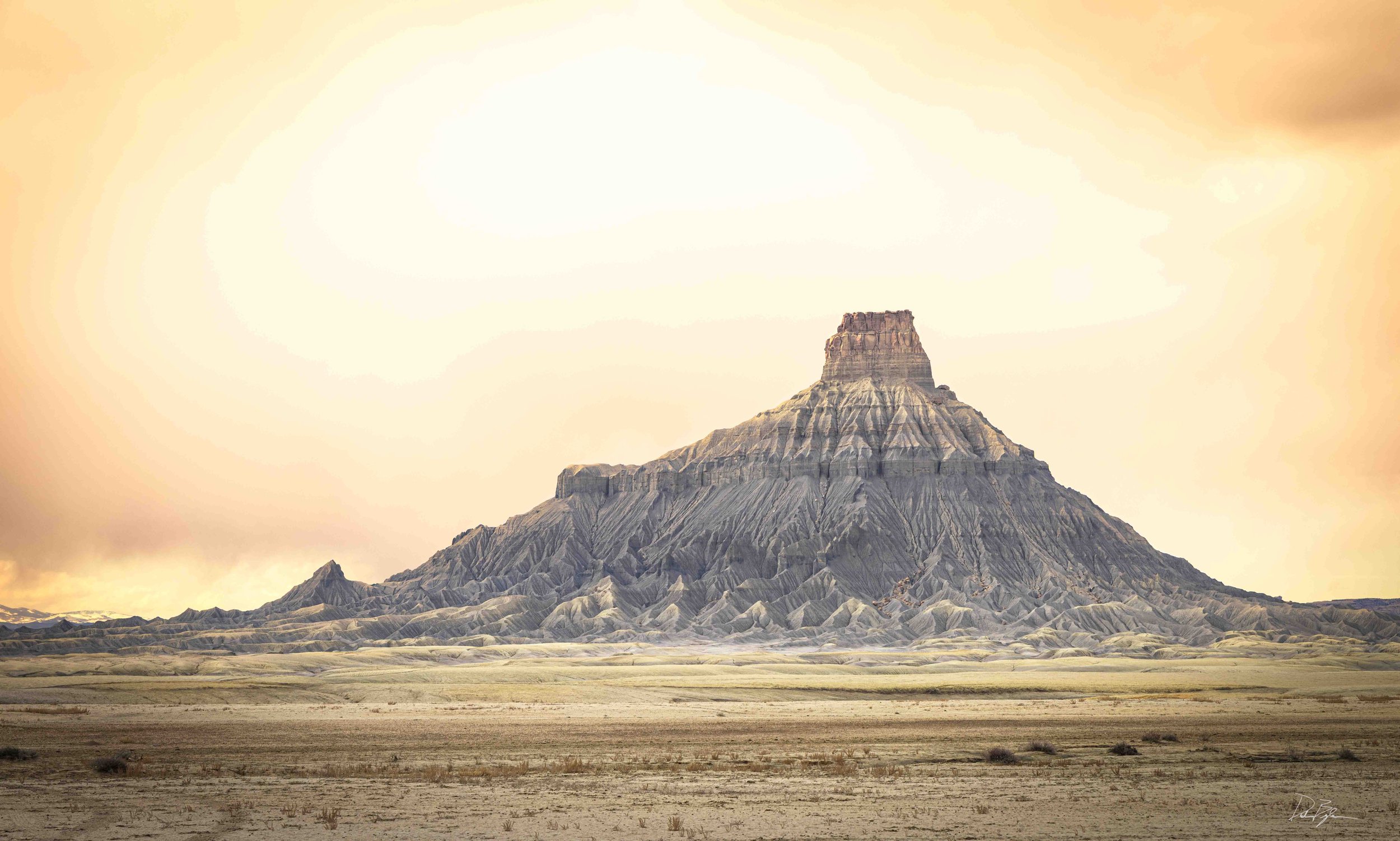 A large mountain with layered rock formations, likely a butte, set in a dry desert landscape with sparse vegetation, under a sky with a warm, hazy glow.