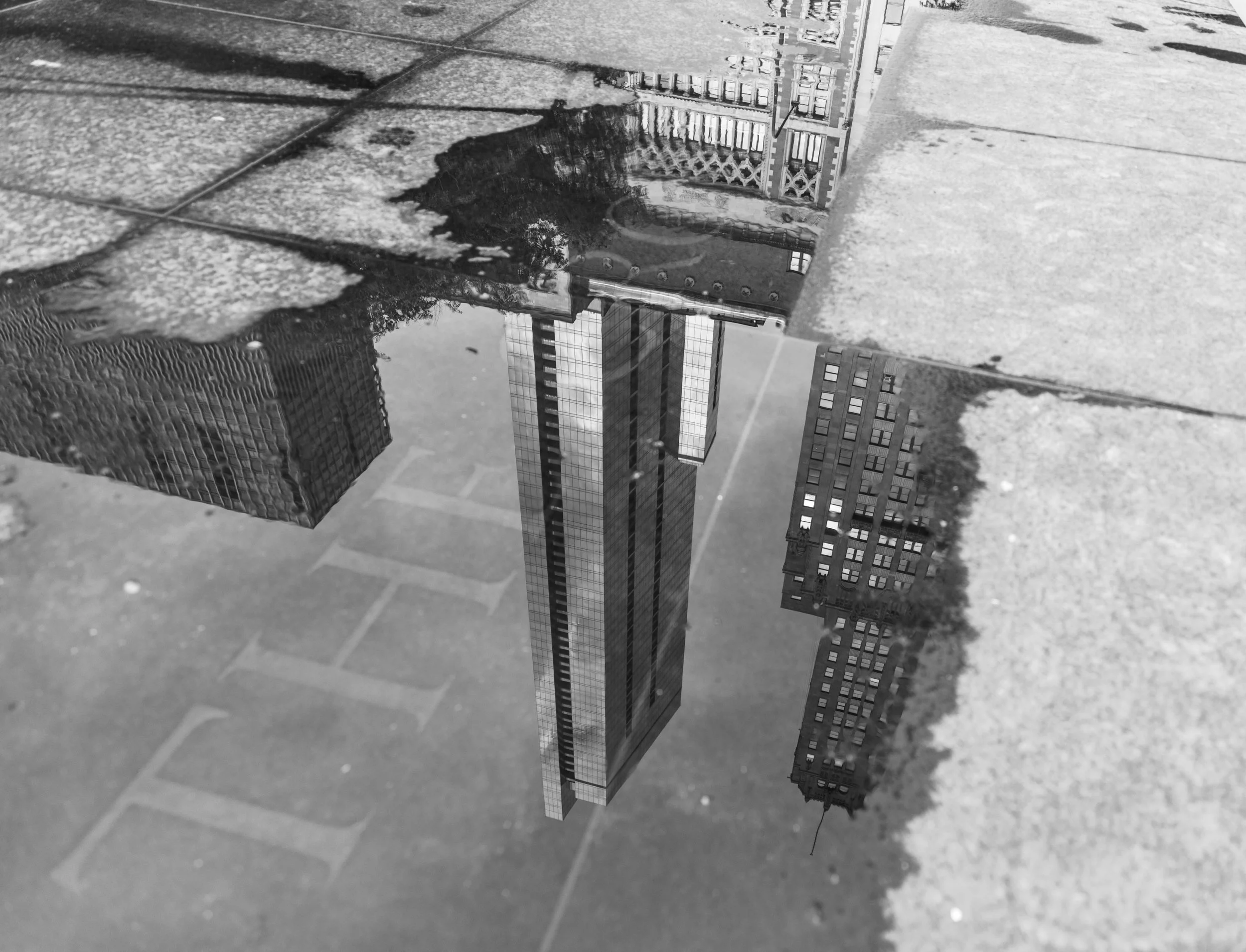Black and white photo of city buildings reflected in a puddle on the pavement.