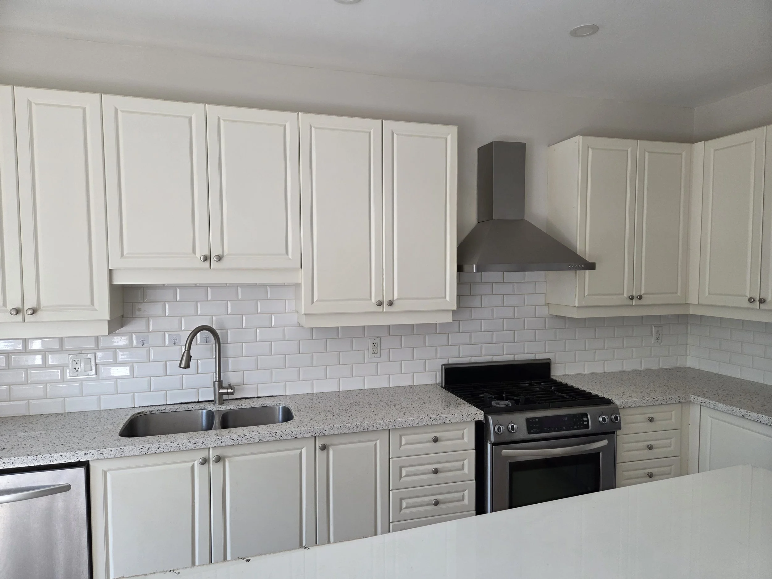 White kitchen cabinets with silver knobs, stainless steel vent hood, white subway tile backsplash, gray speckled countertop, stainless steel sink with a chrome faucet, black oven and stove with digital controls, and multiple electrical outlets along the backsplash.