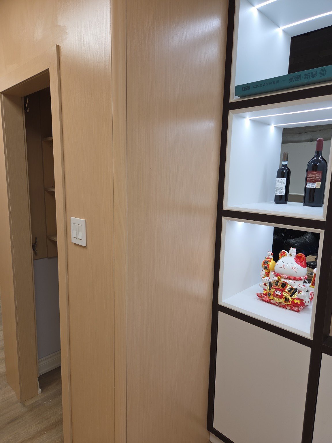 Interior view of a room showing a light wood sliding door closet, a white light switch, and a black shelving unit with illuminated display boxes. The display contains two bottles of wine and a maneki-neko figurine.