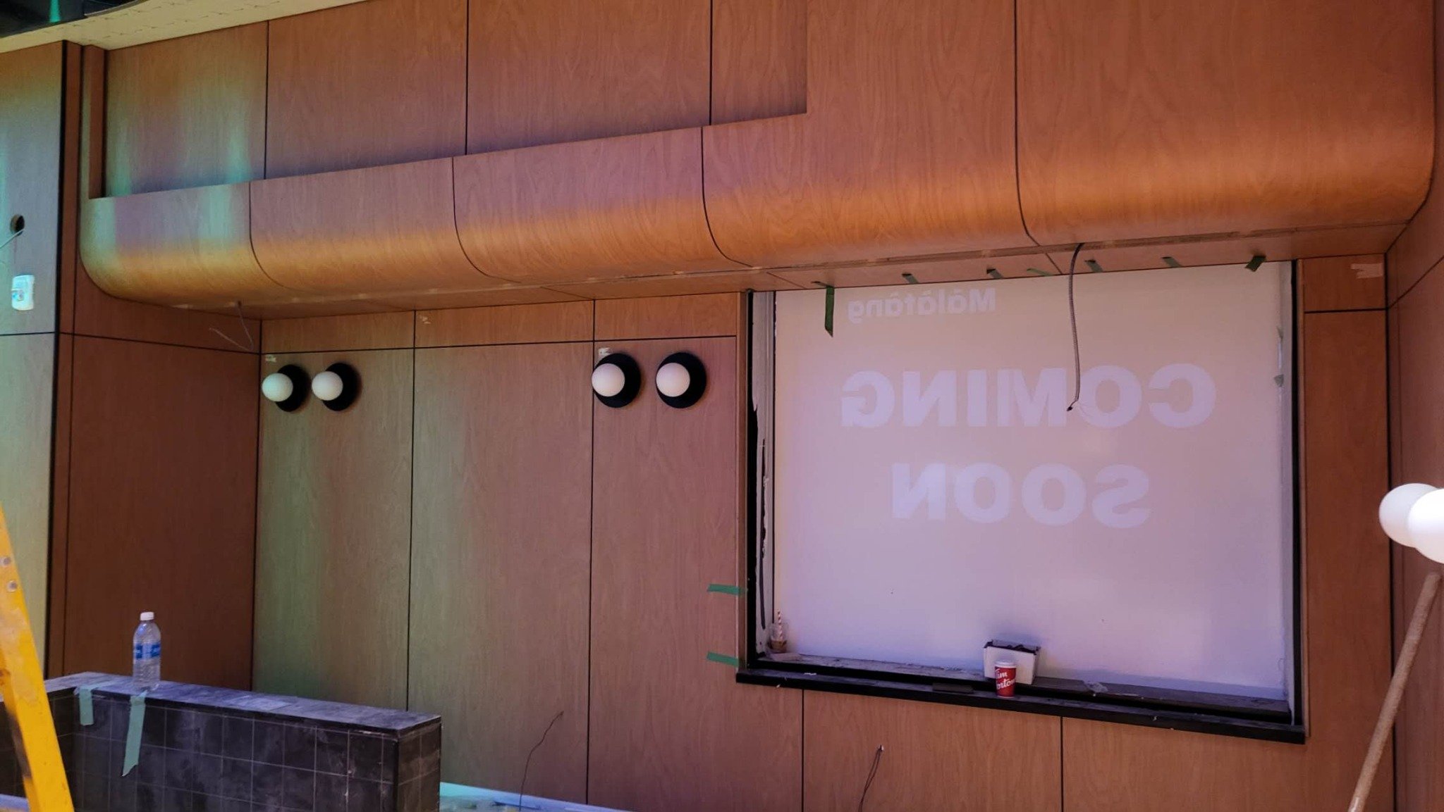 A room under renovation with wooden wall panels, a whiteboard with partially visible text, and construction materials such as a yellow ladder, a water bottle, and a small table.