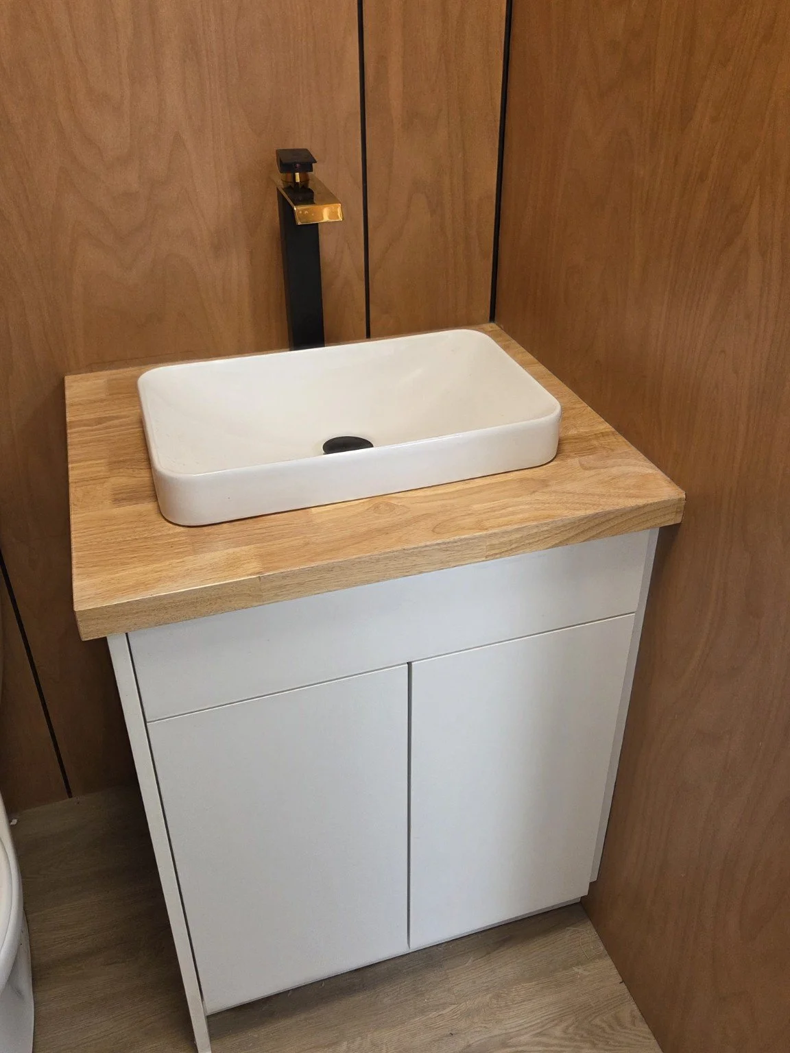 A small white sink with a rectangular basin sits on a wooden countertop, with a sleek black and gold faucet mounted on the wall behind it.
