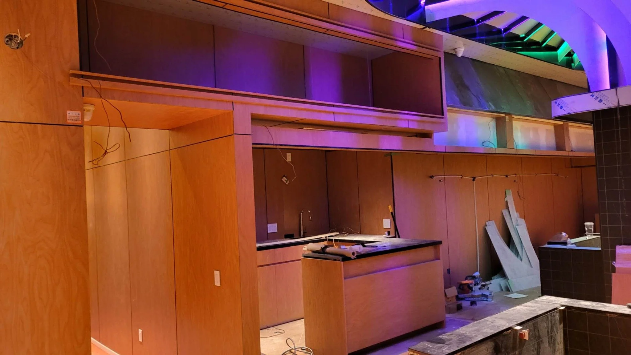 Interior under construction with wooden cabinets, purple and green lighting, and construction materials scattered on the floor.