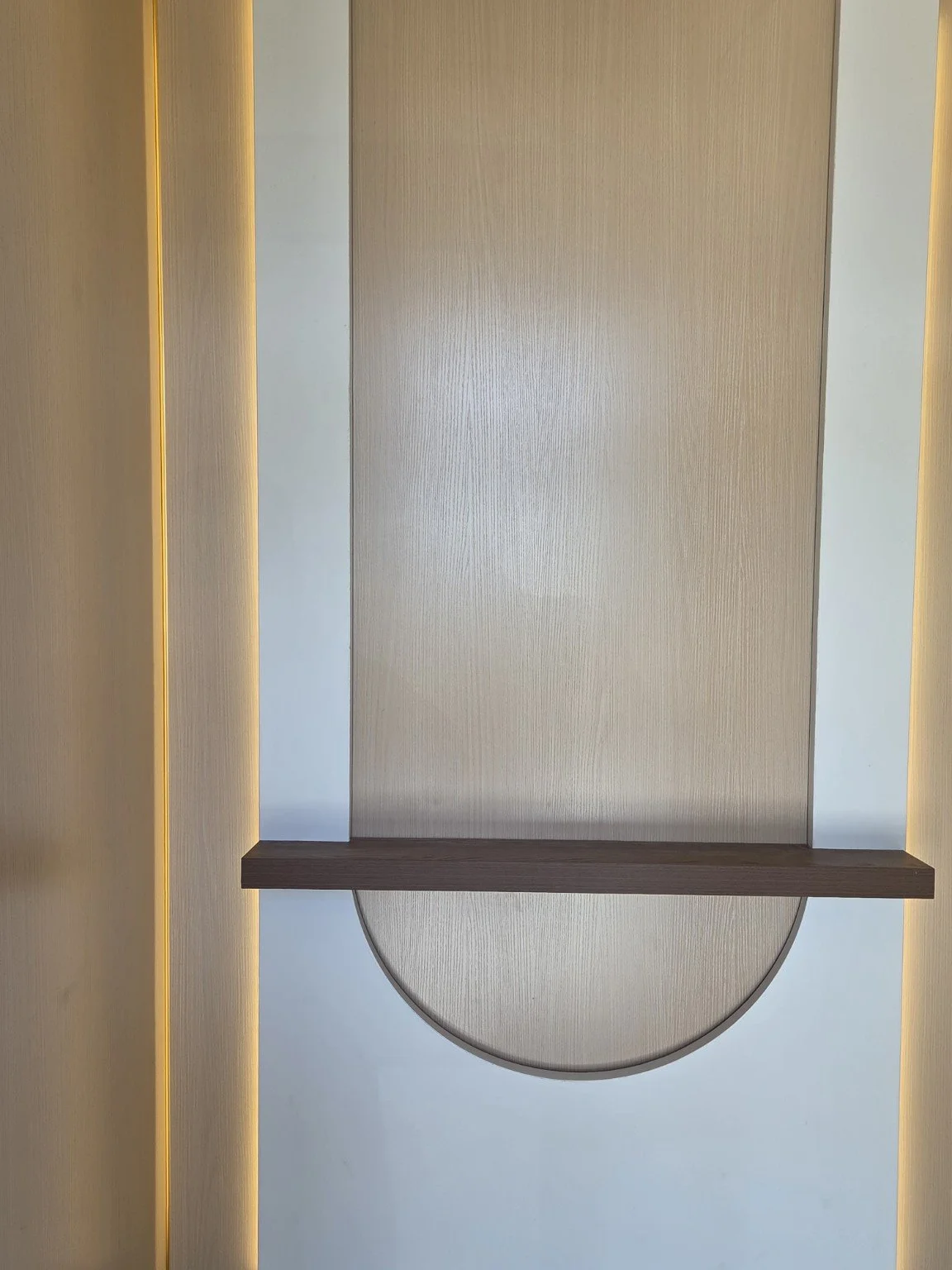A modern interior wall with a large vertical wooden panel, white walls on either side, and a semi-circular design element at the bottom, illuminated by yellow LED lighting.