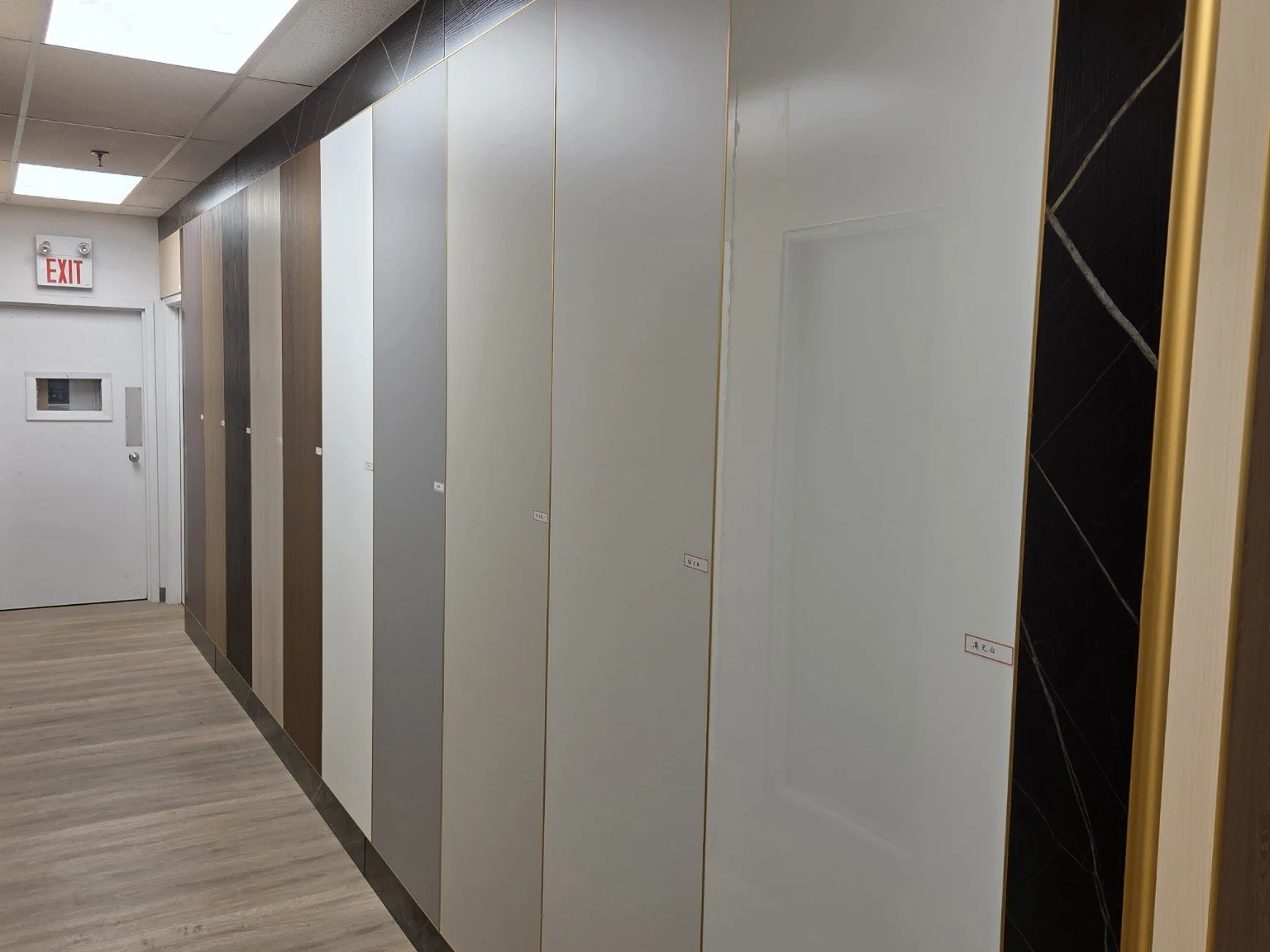 A display of various wall panels in a showroom, featuring different colors and textures with small labels, next to a white door with a window and an exit sign overhead.