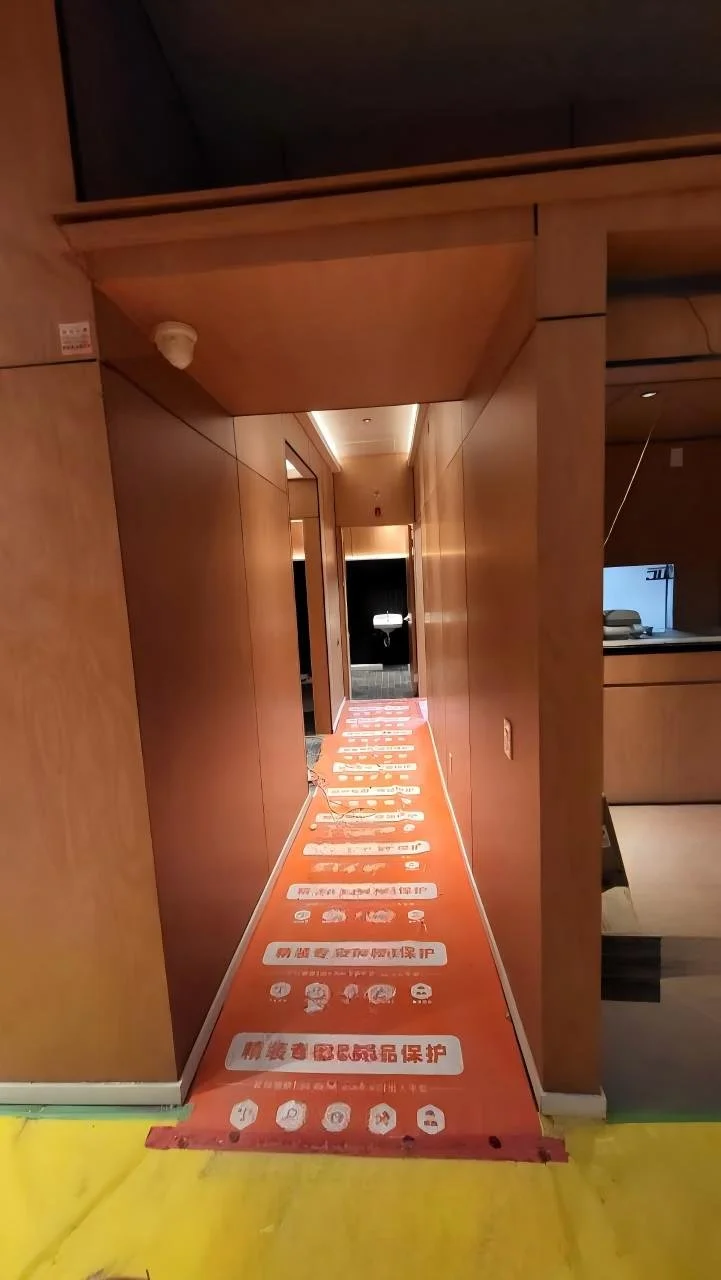 Corridor with orange safety mat and Chinese text, leading to a bathroom with a toilet. Wood-paneled walls and ceiling lighting.