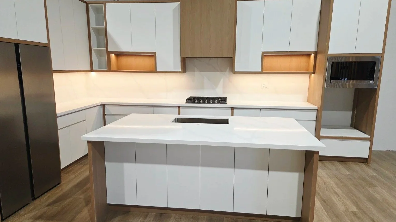 Modern kitchen with white cabinets, a white countertop, and a kitchen island with a sink. Stainless steel appliances include a refrigerator on the left and a microwave on the right. The backsplash is light-colored, and wood accents are visible on the upper cabinets and surrounding the microwave.