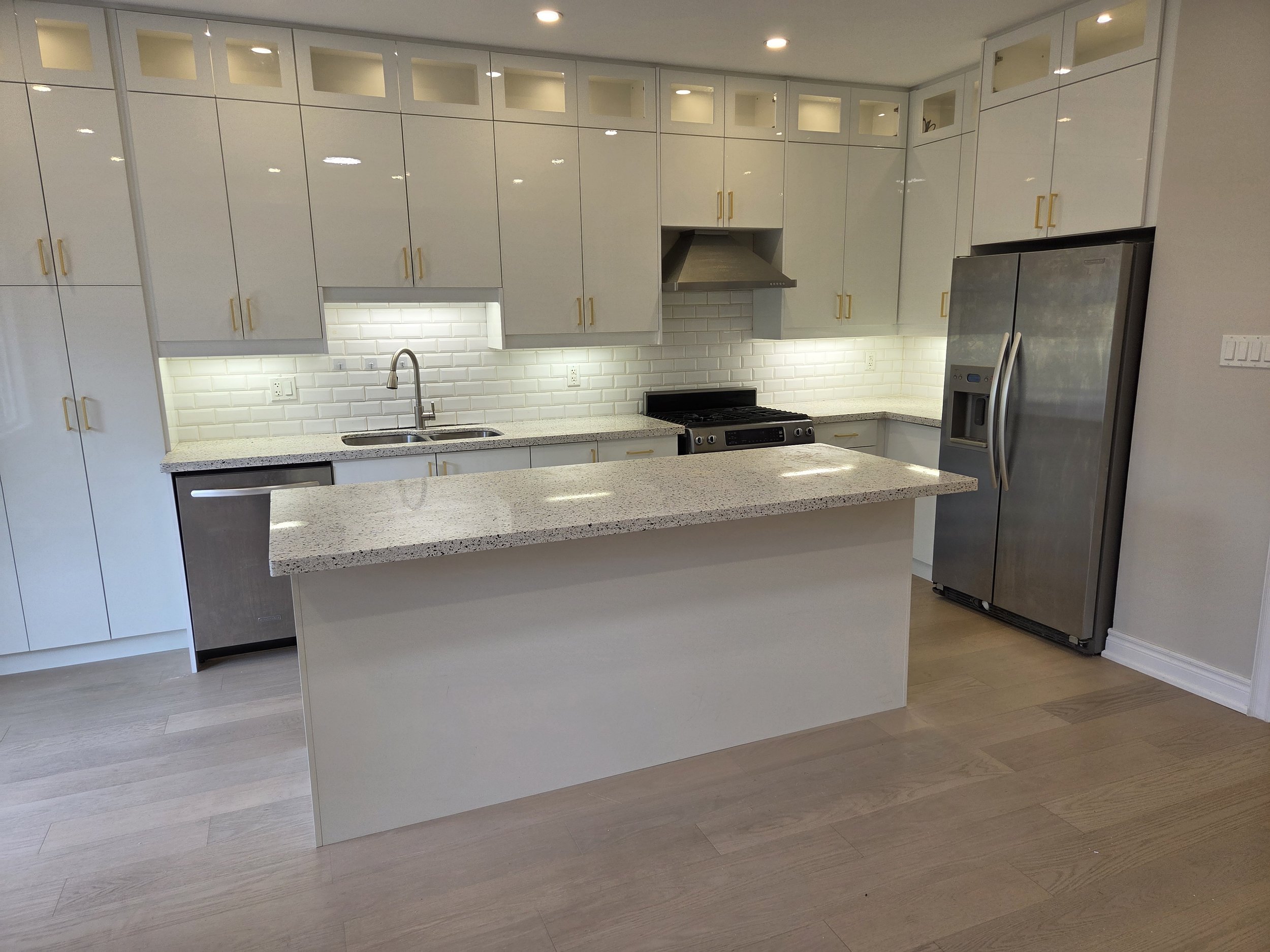 Modern white kitchen with upper cabinets featuring glass inserts, a white subway tile backsplash, granite countertops, stainless steel appliances including a refrigerator and range, and a kitchen island with a speckled surface.