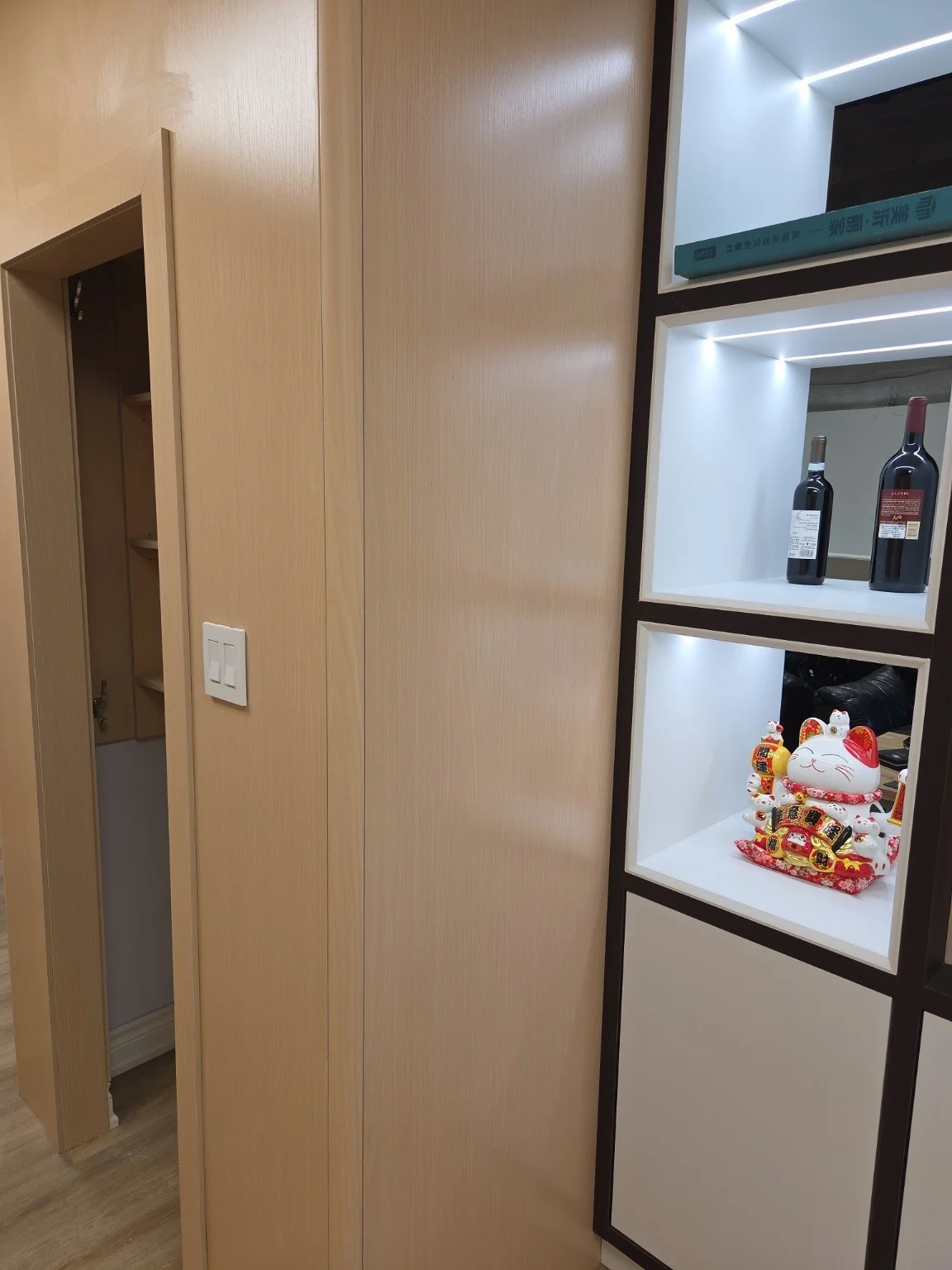 Interior view of a modern living space featuring light wooden panels and a decorative shelf displaying two bottles of wine and a Maneki-neko figurine, with part of a narrow closet or cabinet on the left.