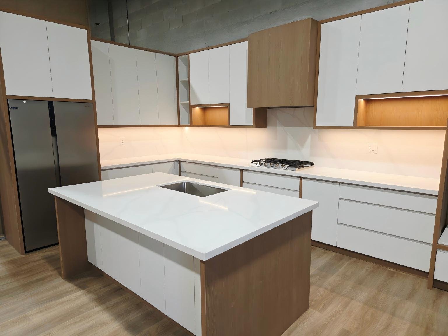 Modern kitchen with white and wood cabinets, a white countertop island with a built-in sink, stainless steel refrigerator, and a gas cooktop, illuminated by under-cabinet lighting.