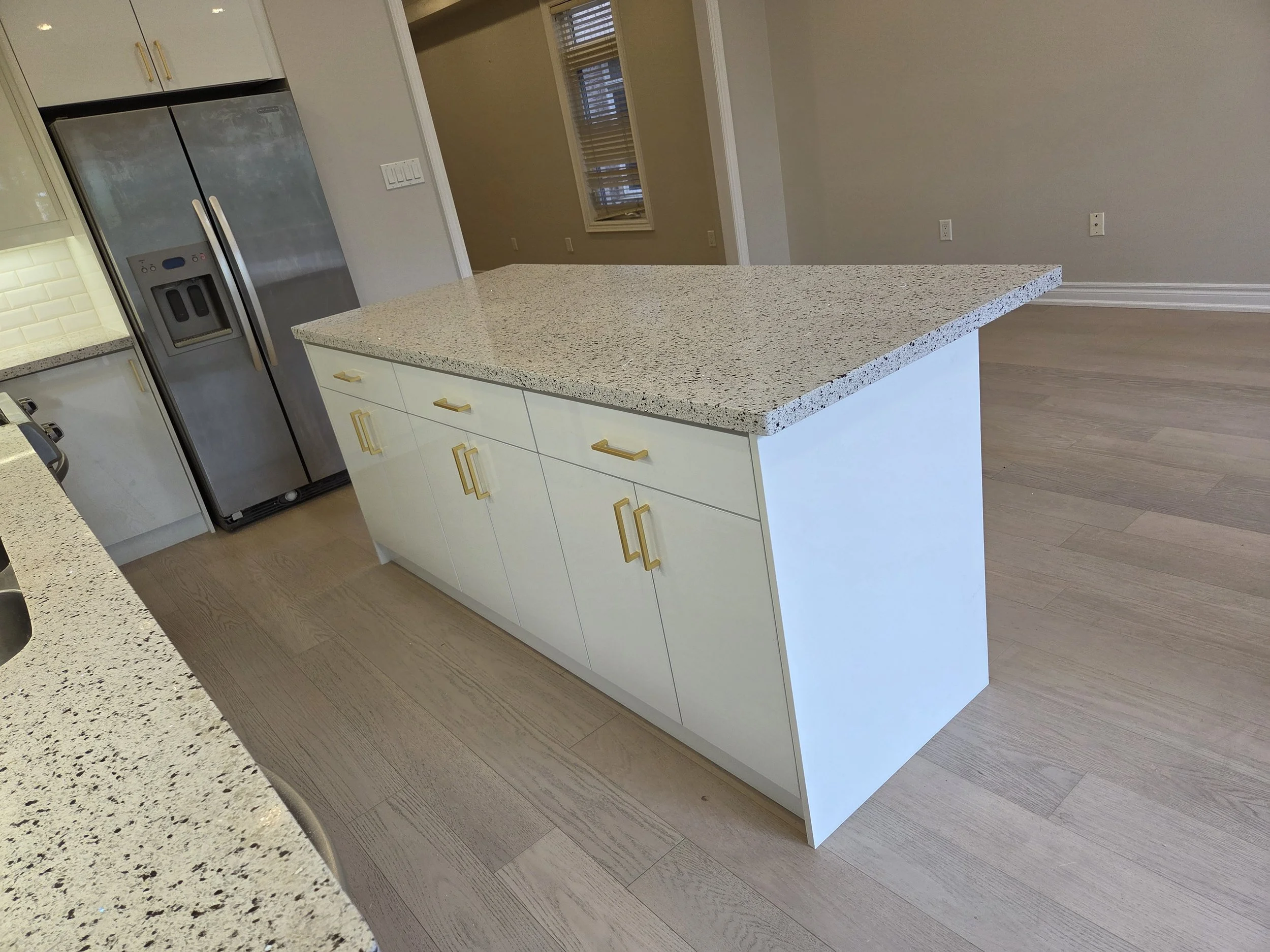 A kitchen island with a speckled granite countertop, white cabinets with gold handles, adjacent to a stainless steel refrigerator, in a modern kitchen with wooden flooring and neutral-colored walls.
