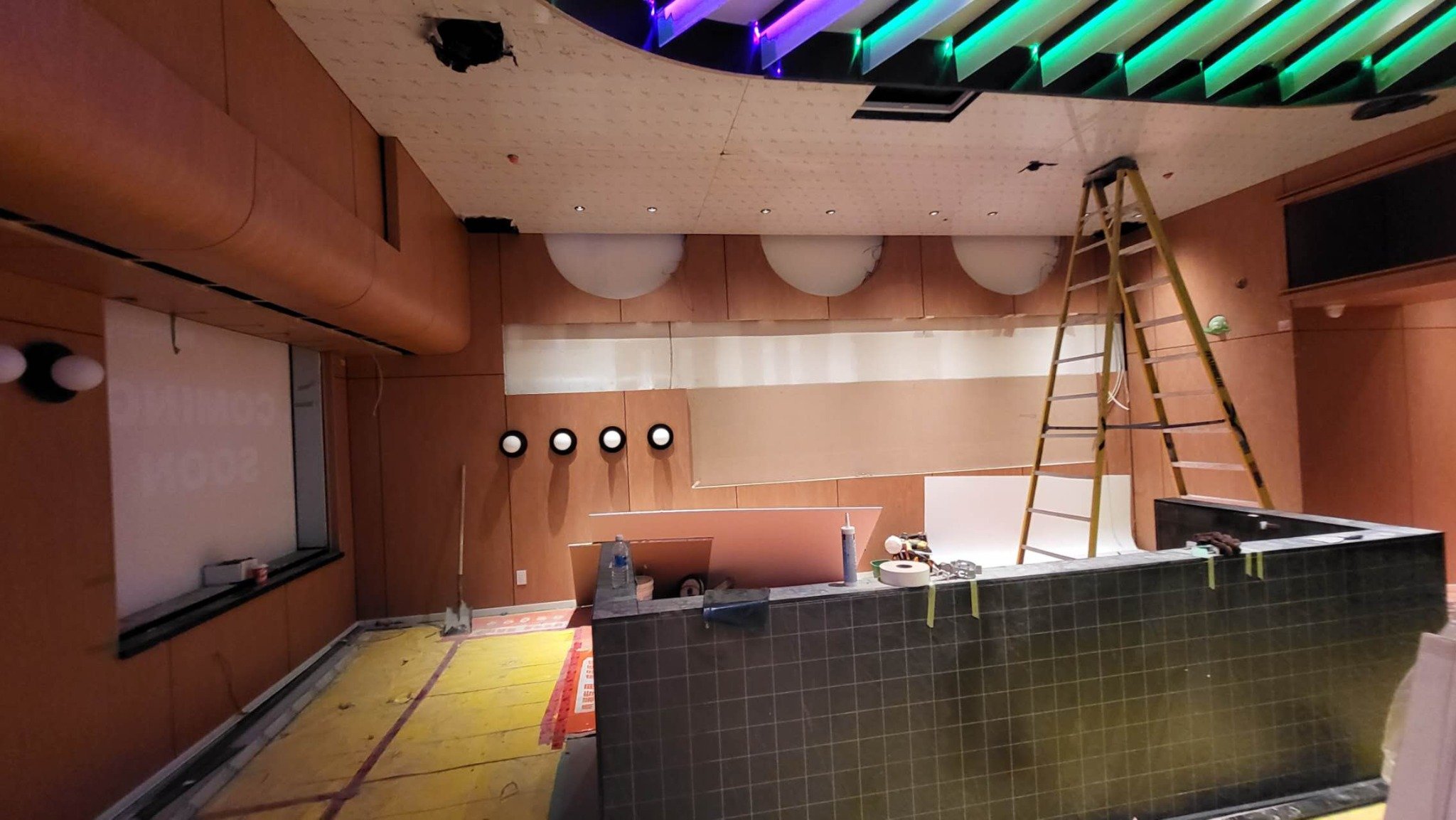 Interior room under renovation with a ladder, construction tools, and lighting fixtures. The ceiling has several holes and exposed areas, and the floor is partially covered with safety mats.