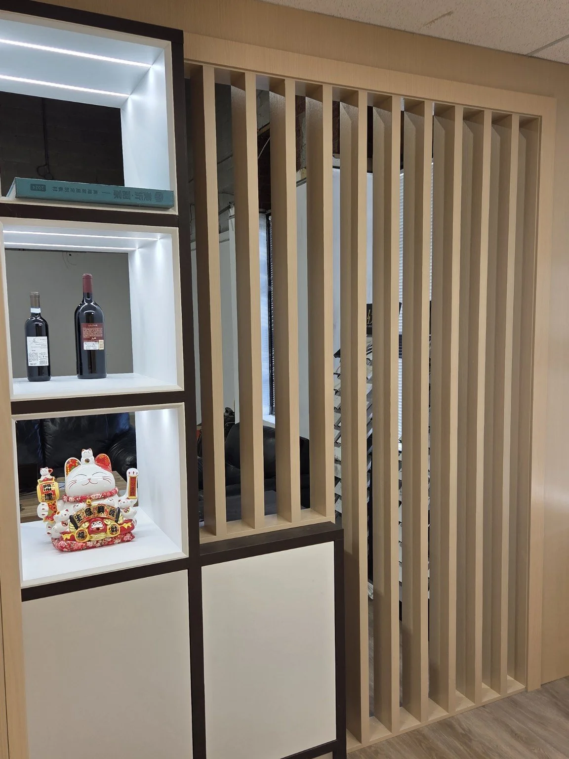 Display shelf with bottles of wine and a Maneki-neko figurine, next to a wooden slat partition in a modern office or restaurant setting.