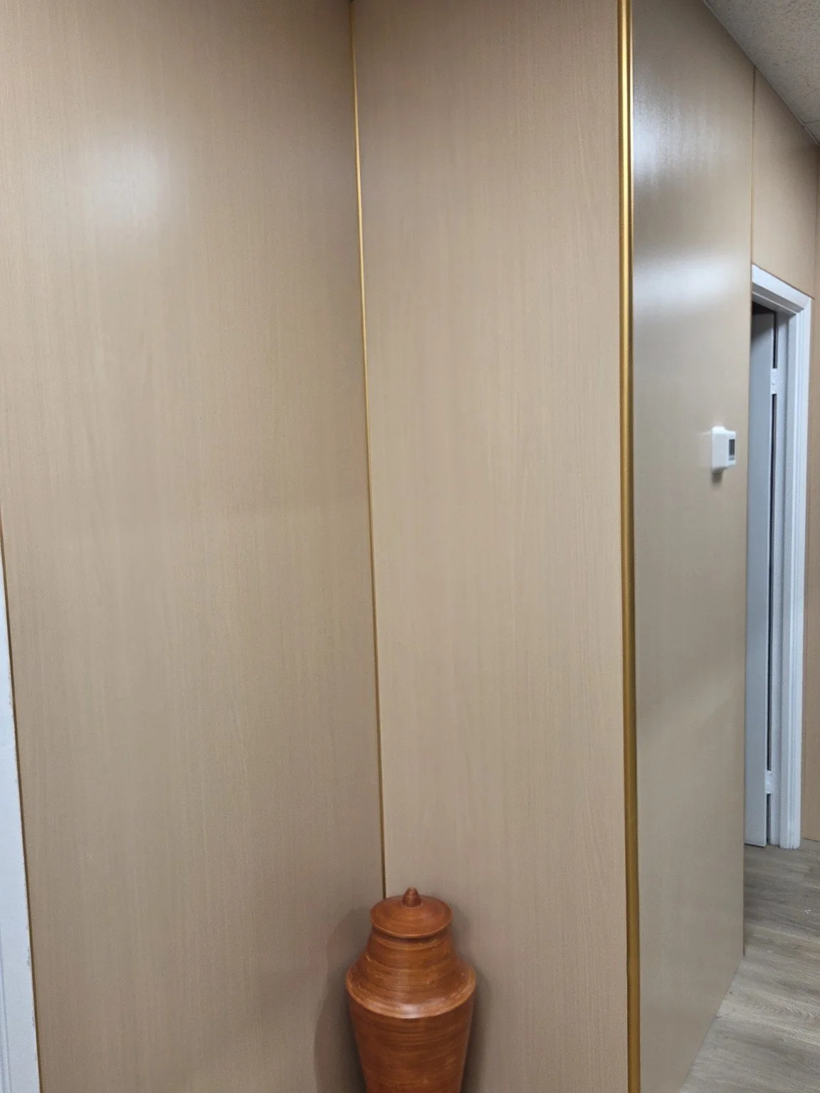 A beige wall with gold vertical trim, a white thermostat on the wall, and a partially open door leading to another room. A large brown ceramic vase sits on the floor in the corner.