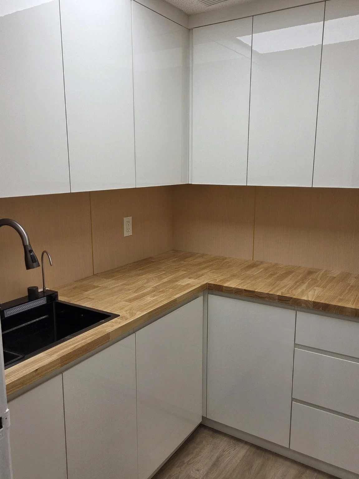 Empty kitchen with white cabinets, wooden countertop, black sink, and beige walls.