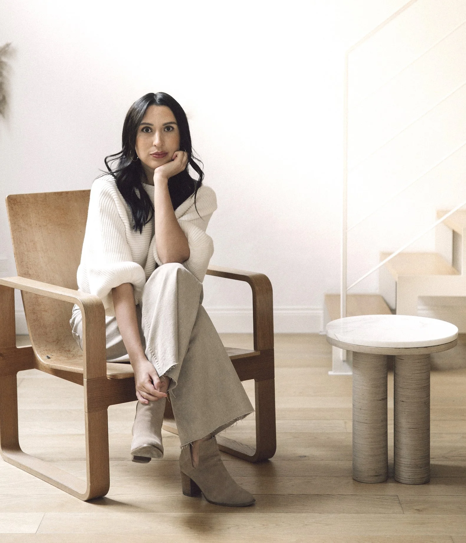 A woman sitting on a wooden chair with her chin resting on her hand, wearing a cream-colored sweater, beige pants, and taupe ankle boots, in a minimalistic room with light wood flooring, white walls, a side table, and a staircase.