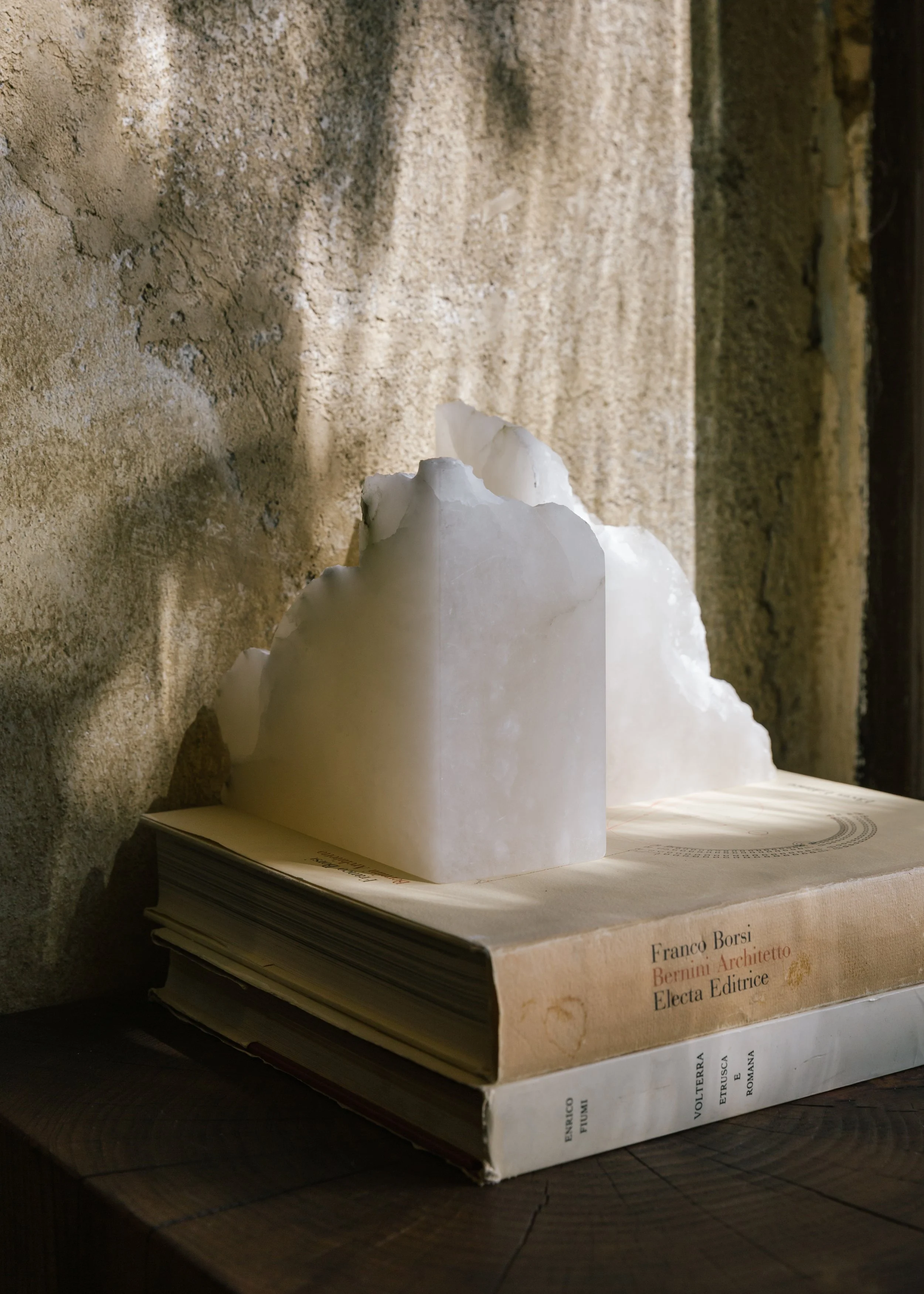 A white marble sculpture of a figure, placed on top of a stack of books, sitting on a wooden surface against a textured wall.