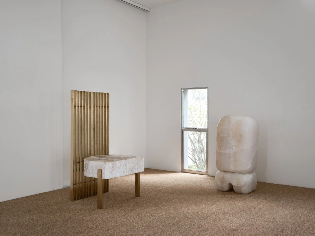 Minimalist interior with a beige textured carpet, a wooden framed bench with light upholstery, a small window with a scene outside, and a large, abstract stone sculpture on the right side of the room.