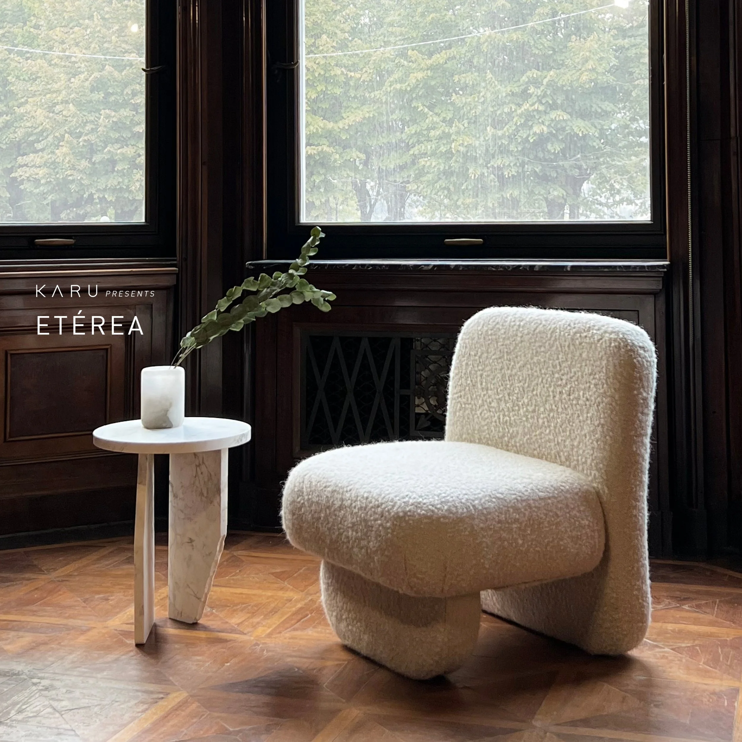 Interior view of a room with a large window showing green trees outside, a white round side table with a white vase containing green foliage, a plush beige armchair, and dark wooden wall paneling.