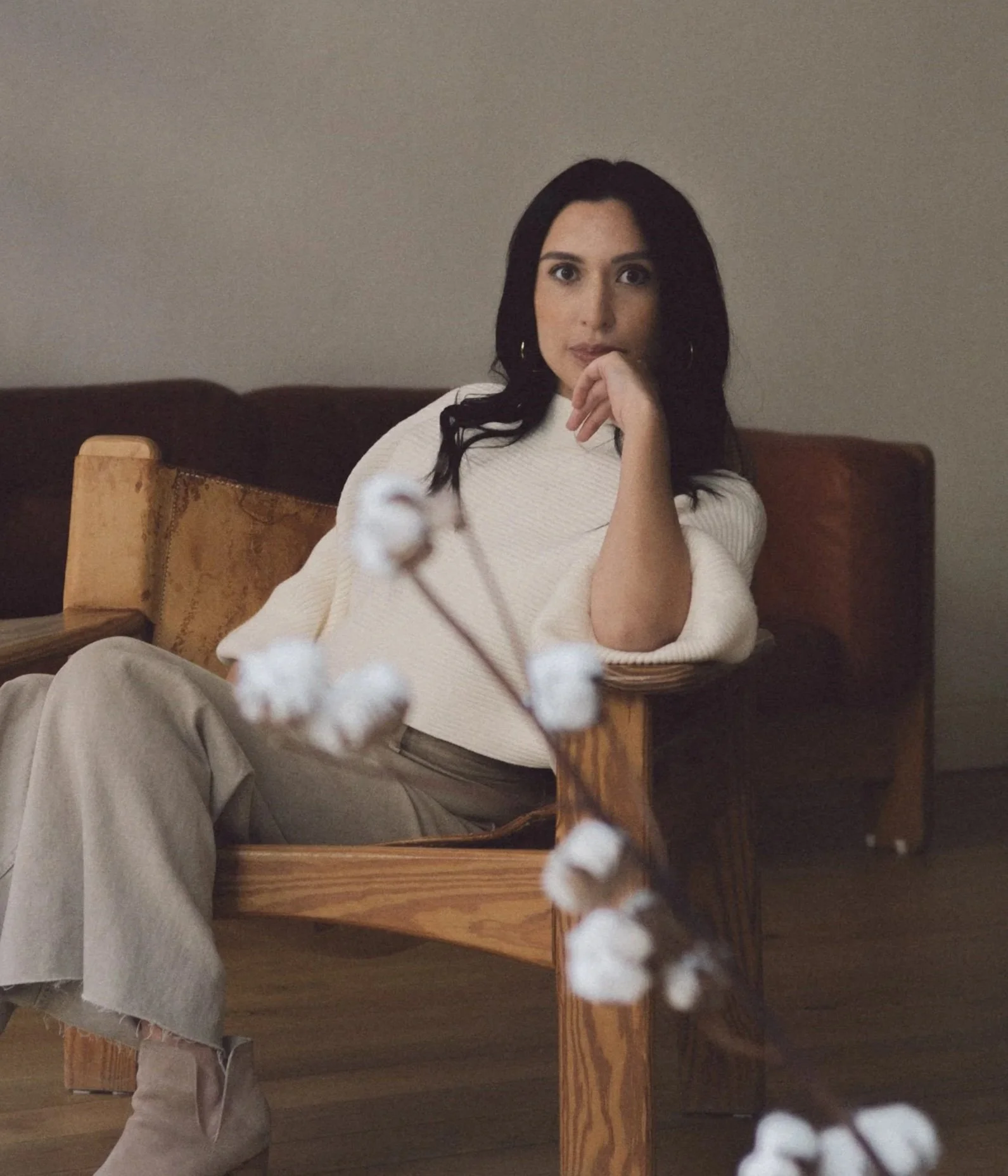 A woman sitting on a wooden chair, wearing a white sweater and beige pants, with dark hair and hoop earrings, in a room with plain walls.