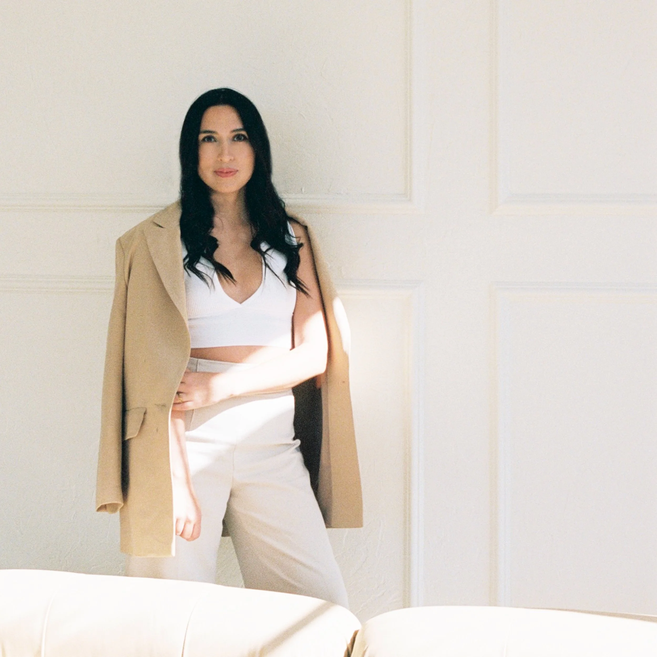 A woman with long black hair, wearing a beige blazer over a white top and beige pants, standing against a plain light-colored wall.