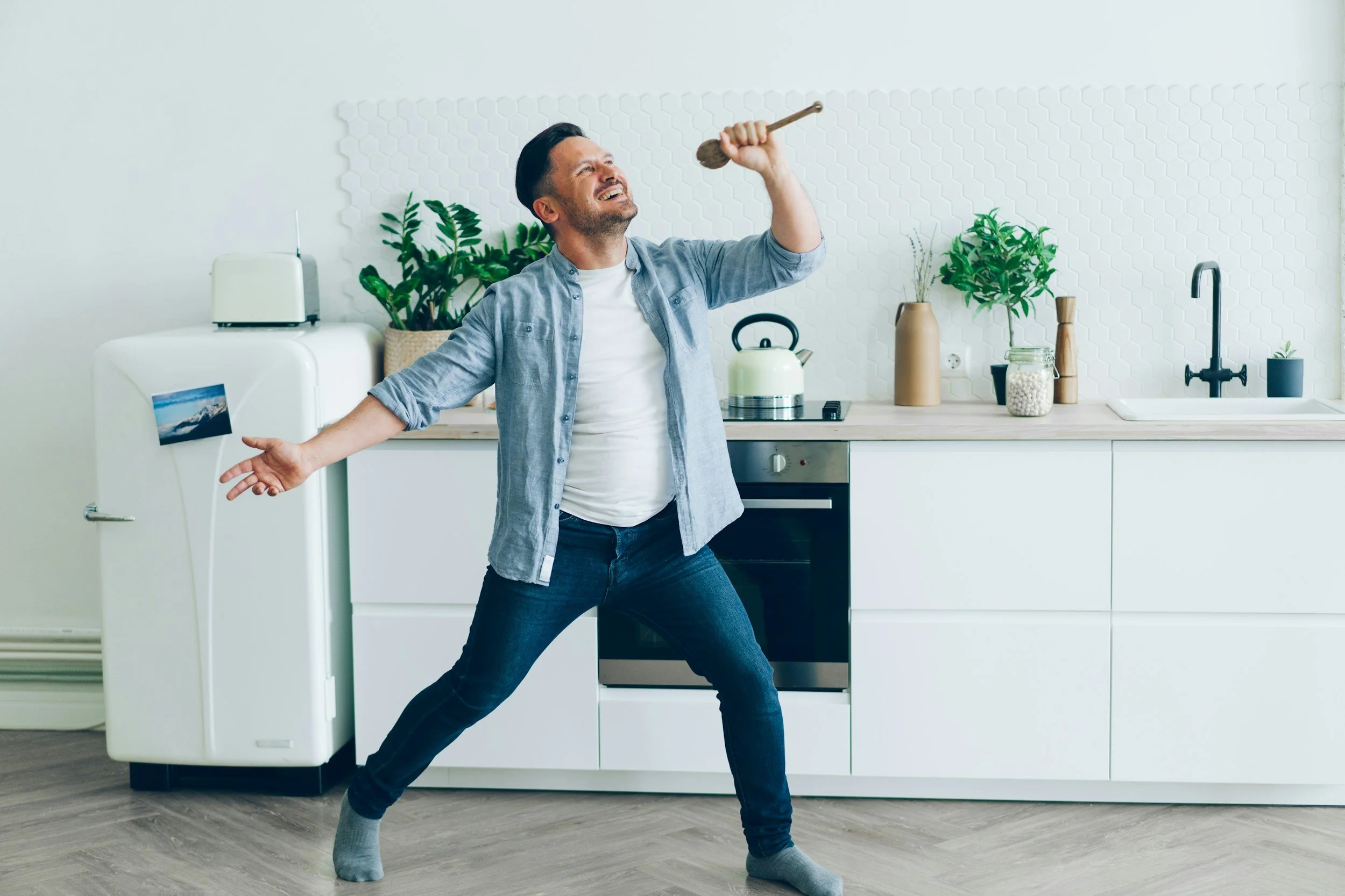 Un homme danse dans une cuisine moderne, tenant une cuillère en bois en l'air avec un grand sourire, portant une chemise en jean et des chaussettes.