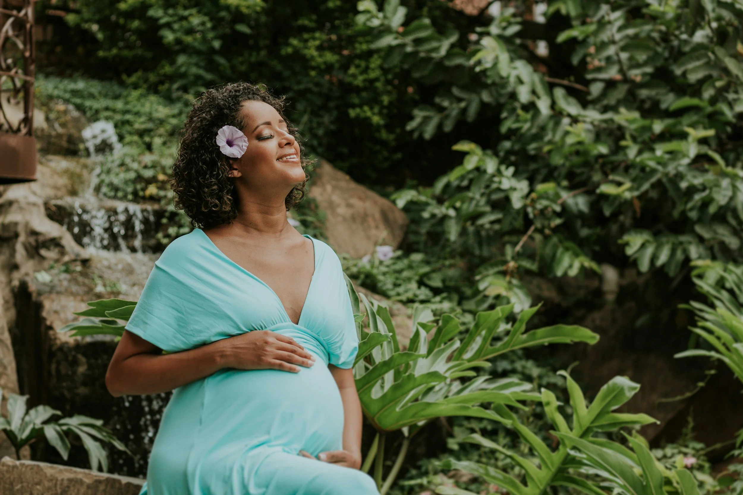Femme enceinte assise dans un jardin verdoyant, portant une robe bleue, avec un hibiscus dans les cheveux, souriant et les yeux fermés.