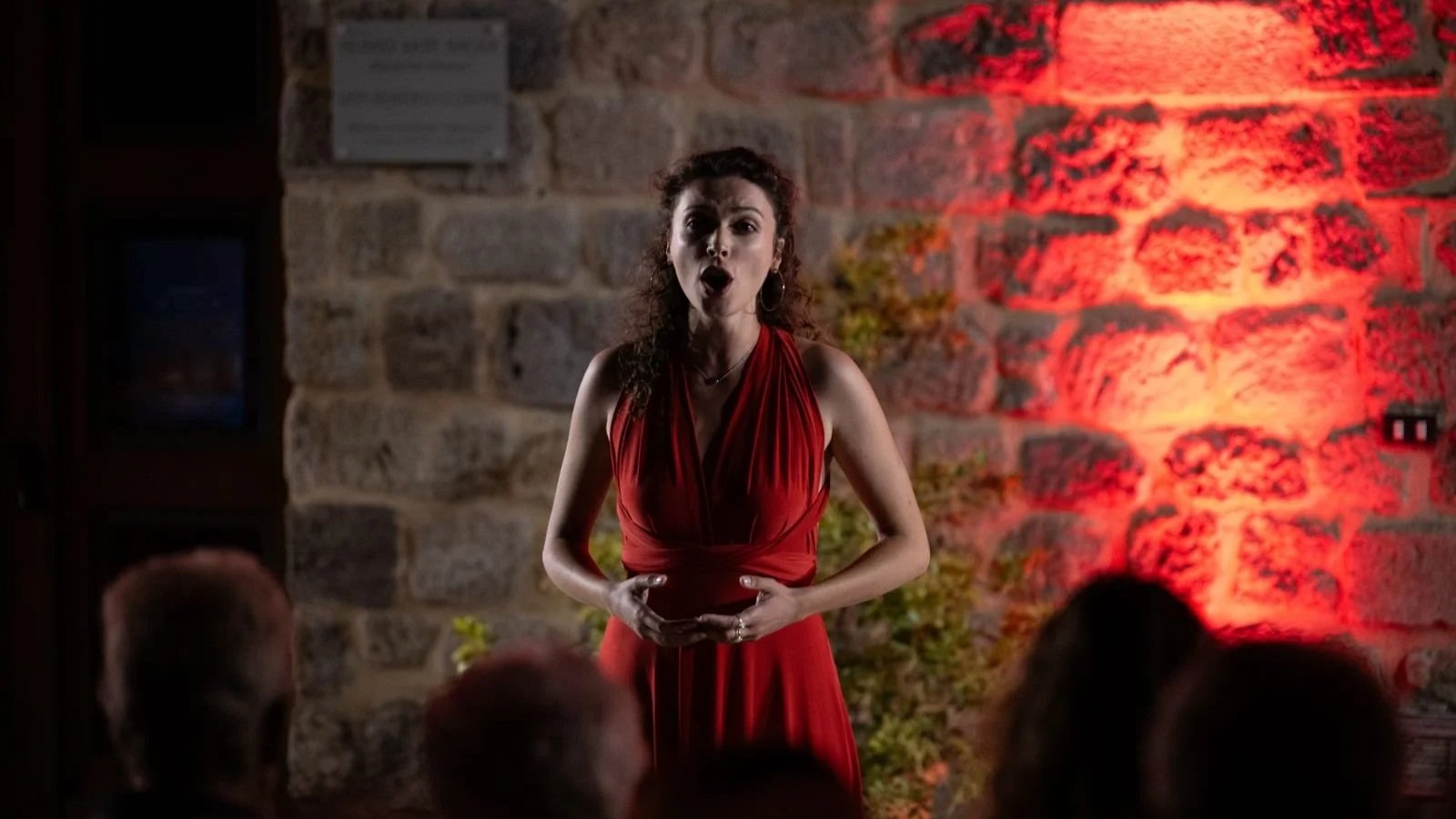 Une femme en robe rouge chante lors d'une performance, face à un public, dans un décor avec un mur en pierre et un éclairage rouge