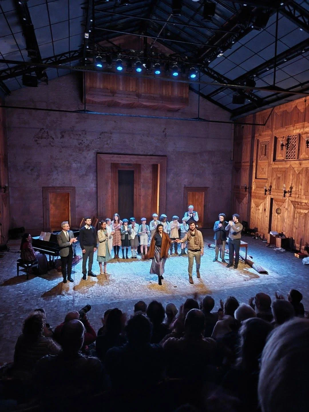 Une scène de théâtre avec plusieurs acteurs sur scène, dont deux au centre se tenant la main, et un groupe d'enfants en arrière-plan. Audience visible en premier plan.