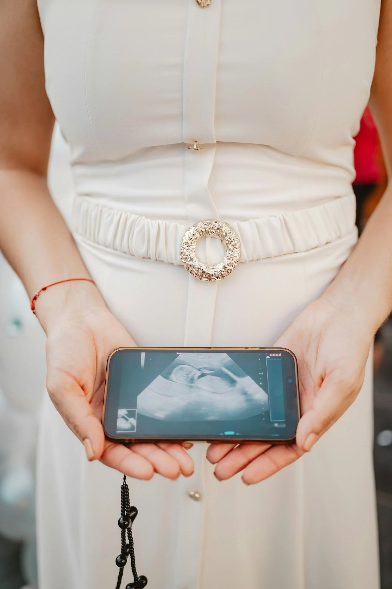 Personne tenant un téléphone portable affichant une échographie, vêtue d'une robe blanche avec une ceinture ornementale et un bracelet rouge.