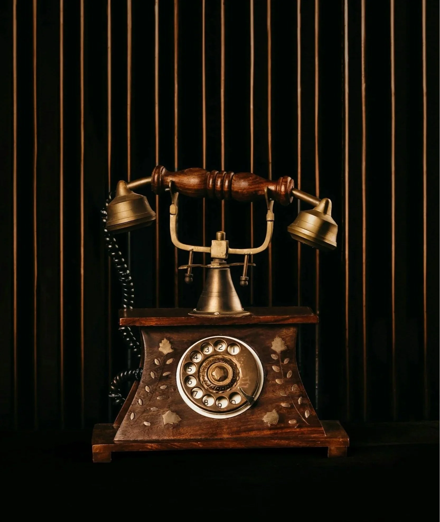 Téléphone ancien en bois et métal posé sur un meuble en bois avec un cadran rotatif, en arrière-plan une grille noire et dorée.
