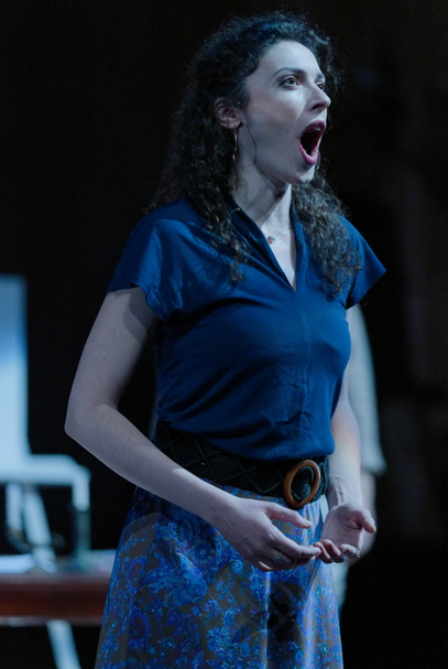 Une femme avec des cheveux bouclés, portant une chemise bleue, chantant ou parlant avec une expression intense.
