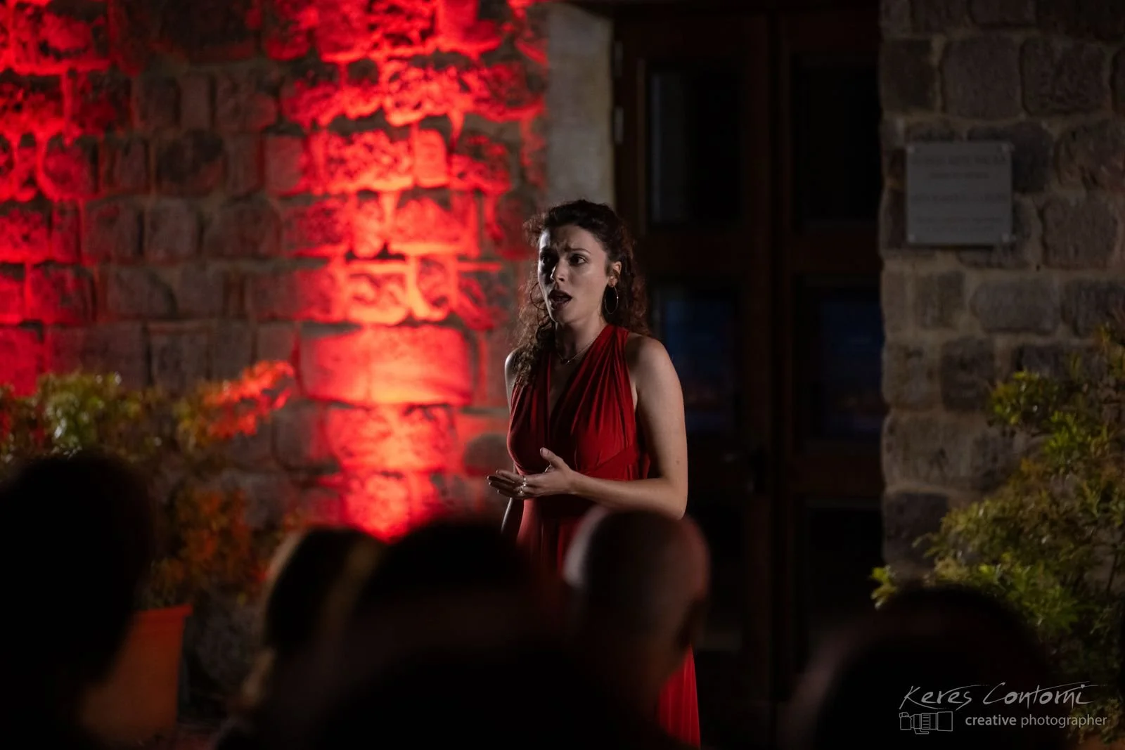 Une femme en robe rouge chante ou parle dans un environnement sombre avec un mur en pierre rouge éclairé en arrière-plan.