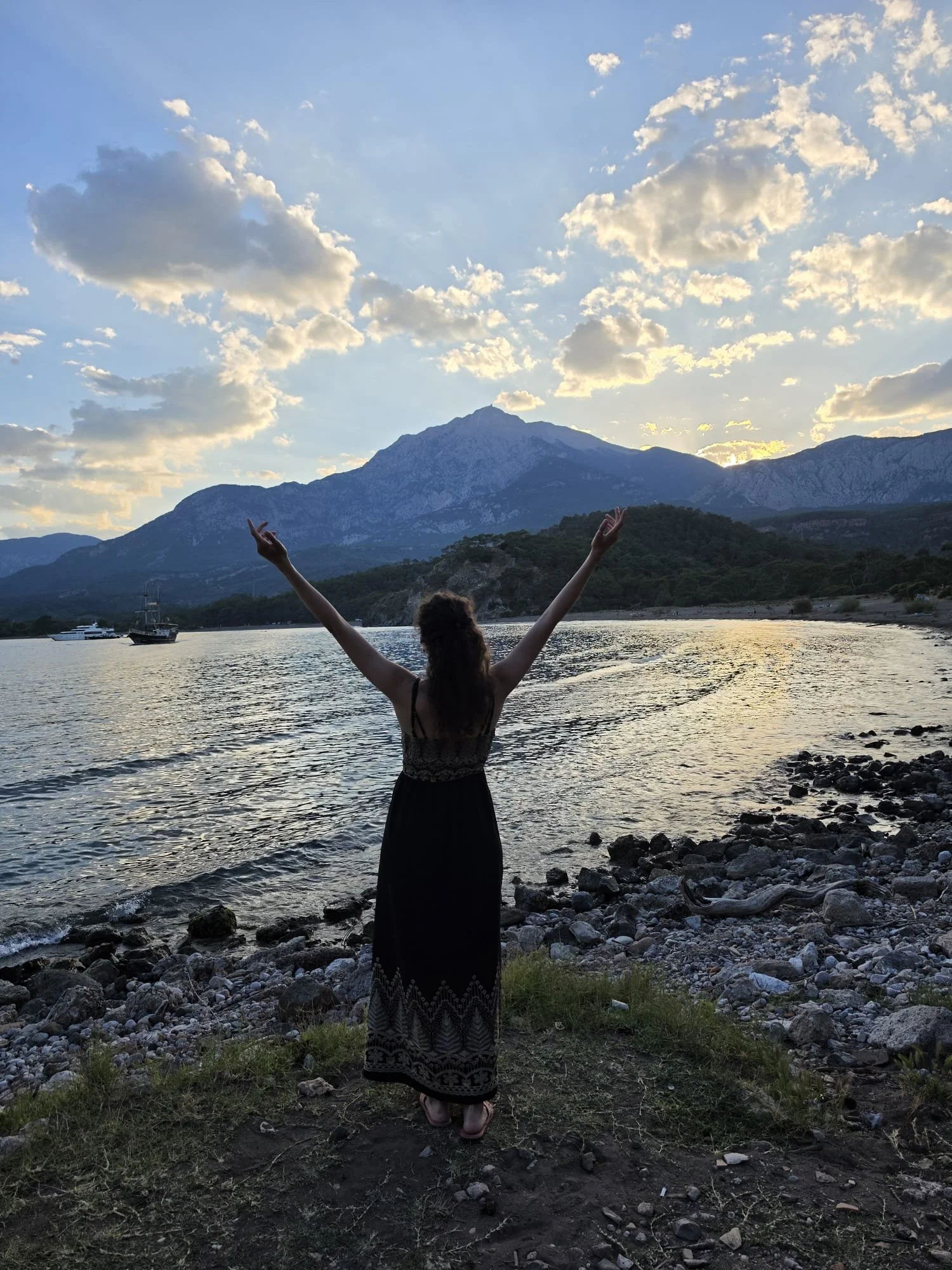 Une femme debout près d'une rivière avec ses bras levés, montagnes en arrière-plan et un ciel avec des nuages au coucher du soleil.