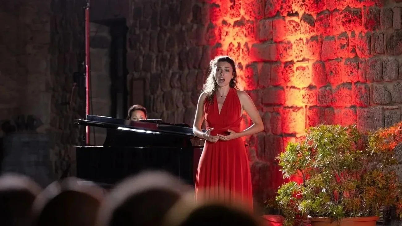 Une femme en robe rouge chante lors d'une performance musicale, avec un piano en arrière-plan et un mur en pierre éclairé en rouge, dans un espace intérieur avec une vigne en pot à droite.