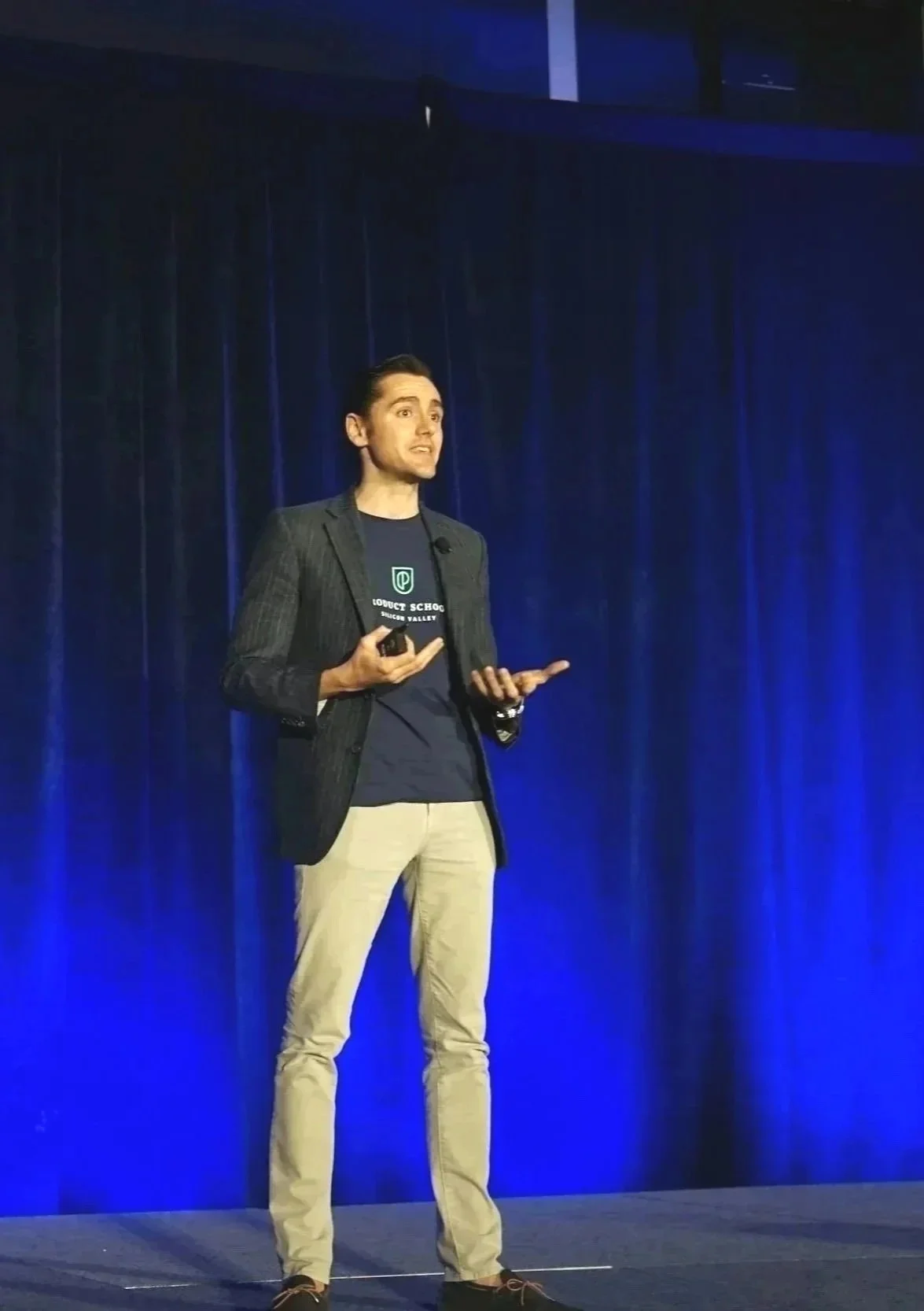 Un homme parle en public lors d'une conférence ou présentation, portant une veste noire, un t-shirt bleu, et des pantalons beige, debout sur scène avec un fond en tissu bleu.
