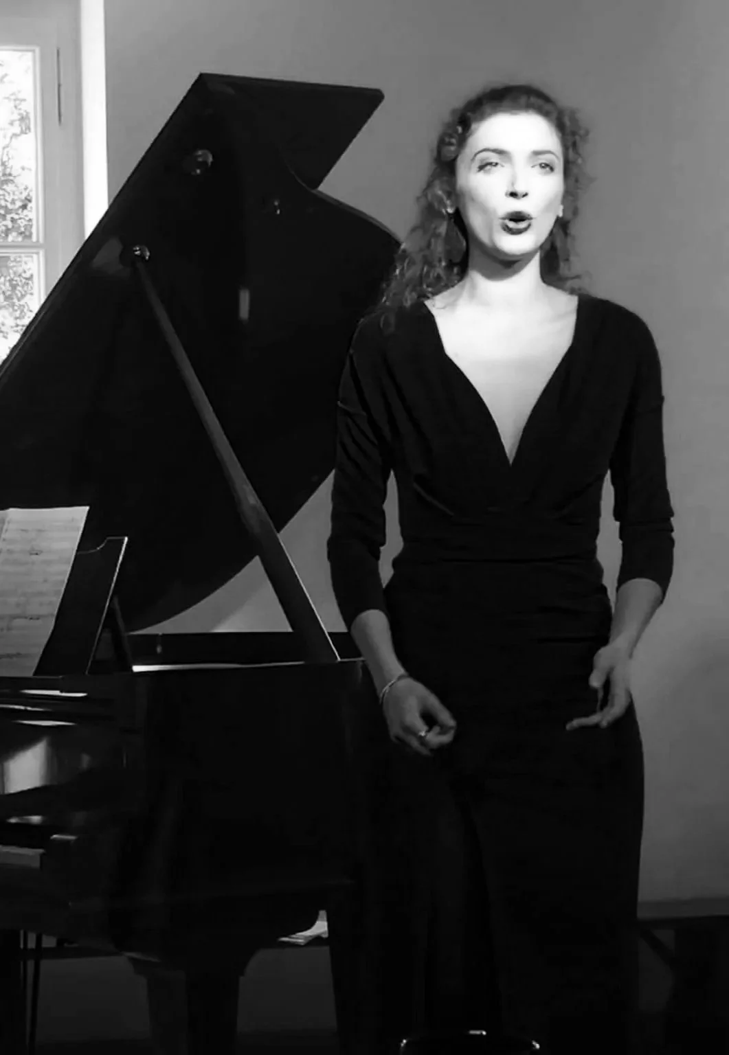 Femme chantant, debout à côté d'un piano à queue, en noir et blanc dans une pièce avec une fenêtre visible.