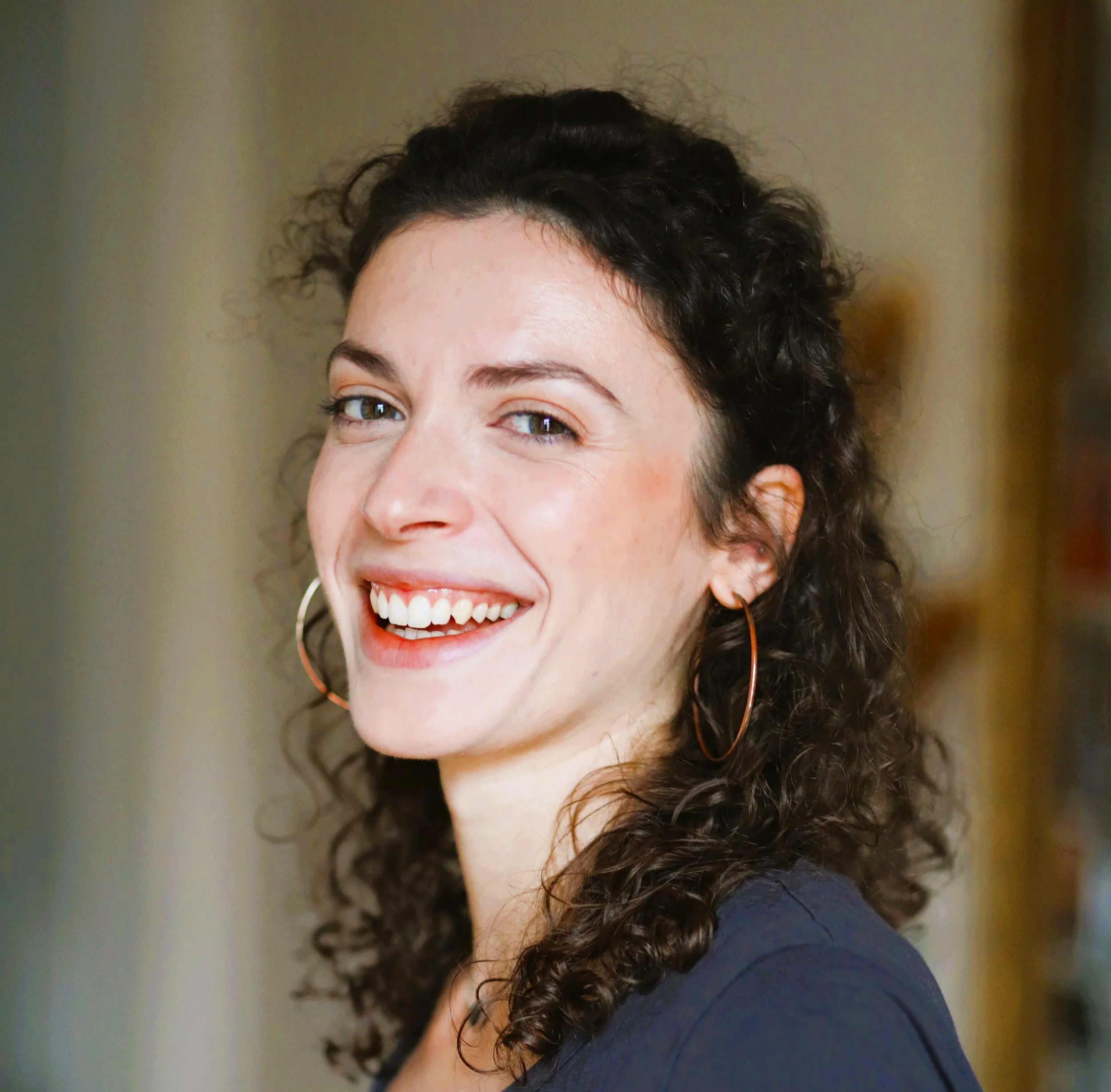 Photo d'une femme avec des cheveux bouclés et un sourire large, portant de grandes boucles d'oreilles en cercle, dans un environnement intérieur.
