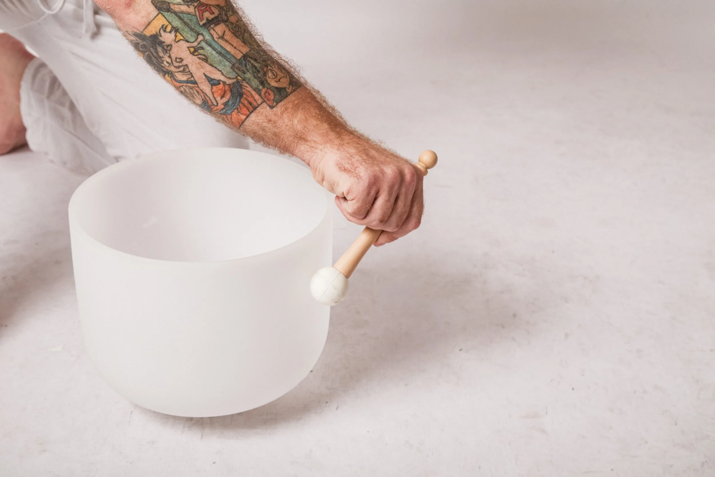 A person with colorful tattoos on their arm is holding a wooden mallet and striking a white singing bowl on a light-colored surface.