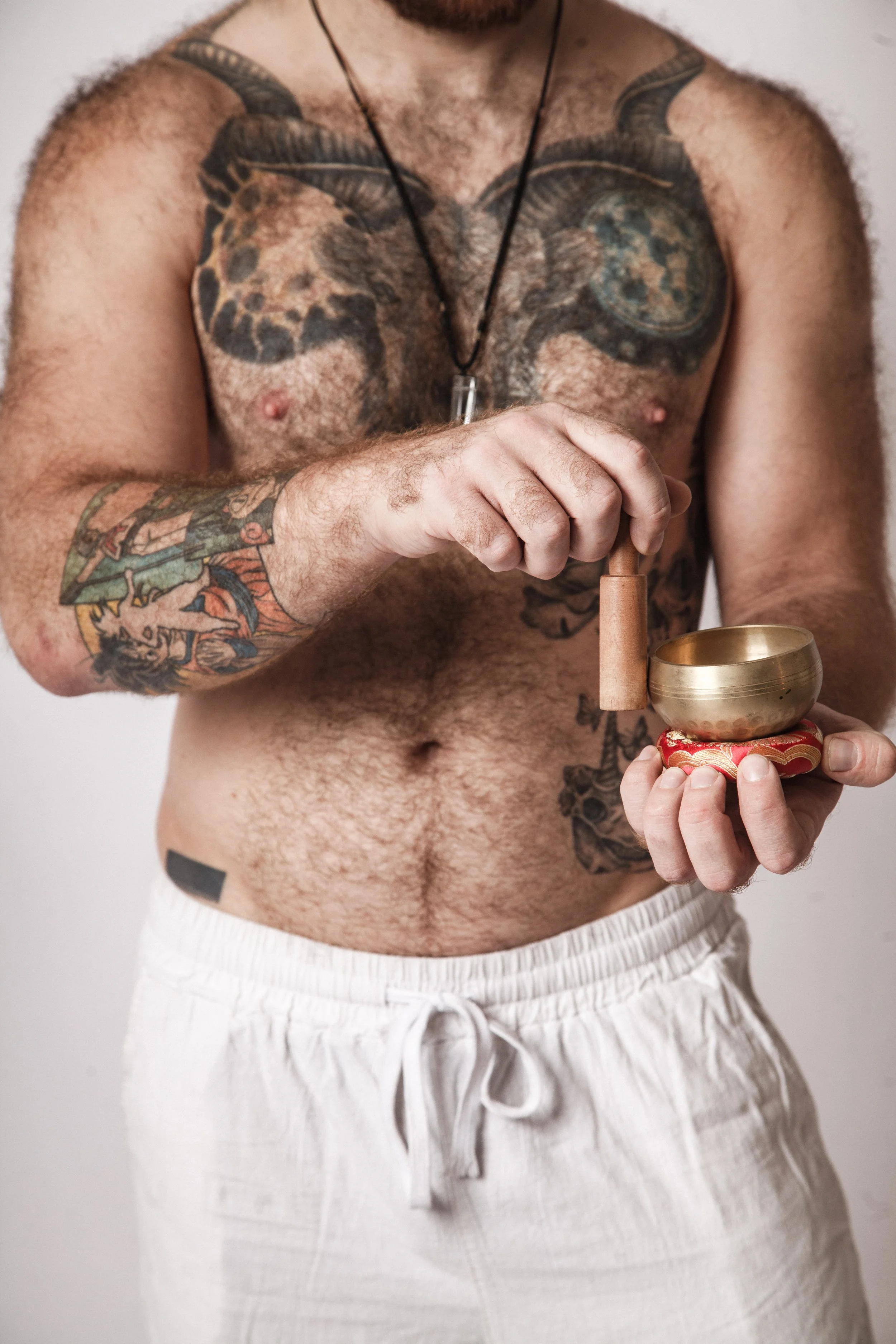 A shirtless man with tattoos on his chest, arms, and shoulder, wearing white shorts and holding a singing bowl with a mallet, standing against a plain background.